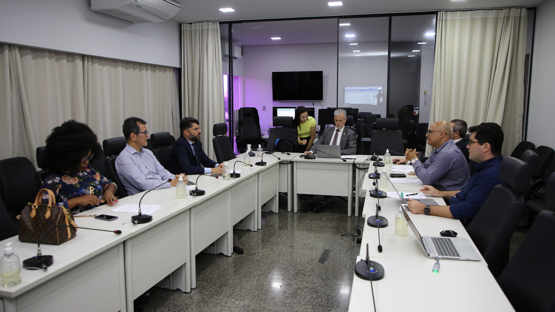 Fotografia colorida onde um grupo de pessoas estão sentados em volta de uma mesa retangular branca com um espaço no meio. Sobre as mesas, estão diversos materiais como notbooks, cadernos e canetas. Eles estão em uma sala branca com duas janelas cobertas de cortinas brancas. Uma das janelas ao fundo está aberta, mostrando outra sala 