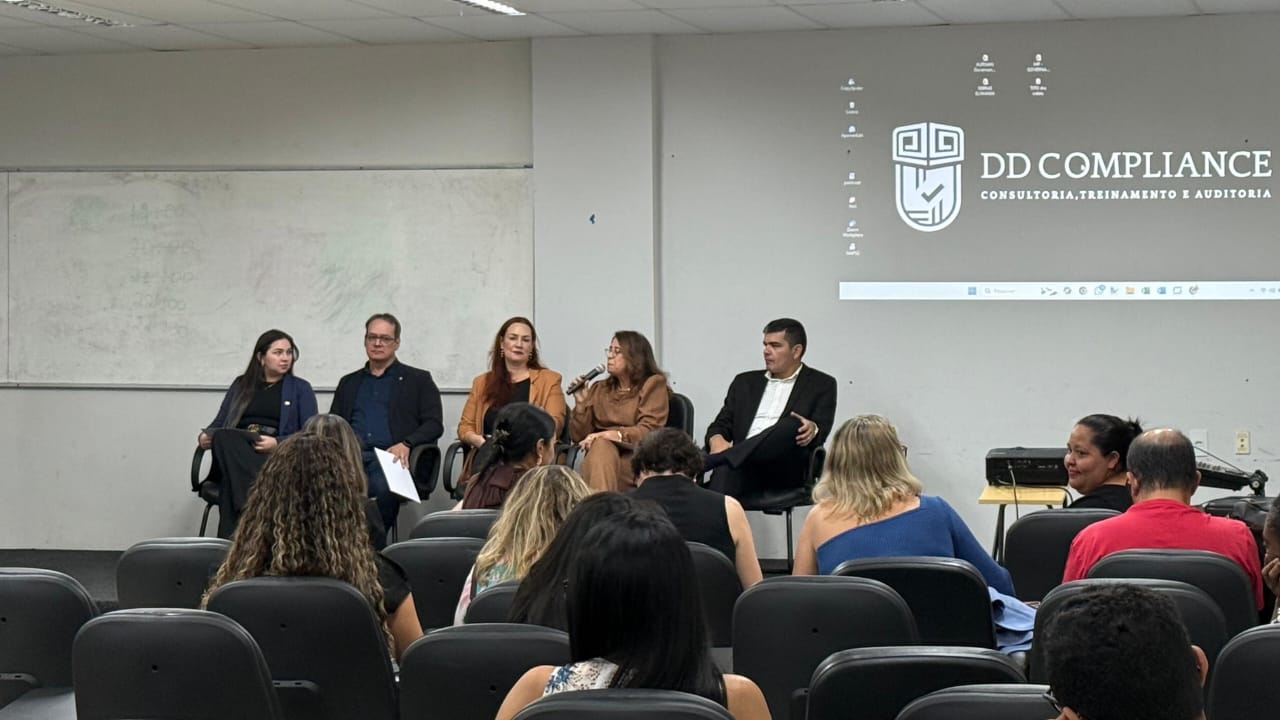 Fotografia colorida onde onde cinco pessoas, três mulheres e dois homens, estão sentados em cima de de um palco enquanto outras várias estão sentadas a frente em cadeiras de borracha escura. Todos estão em uma sala de paredes brancas. 