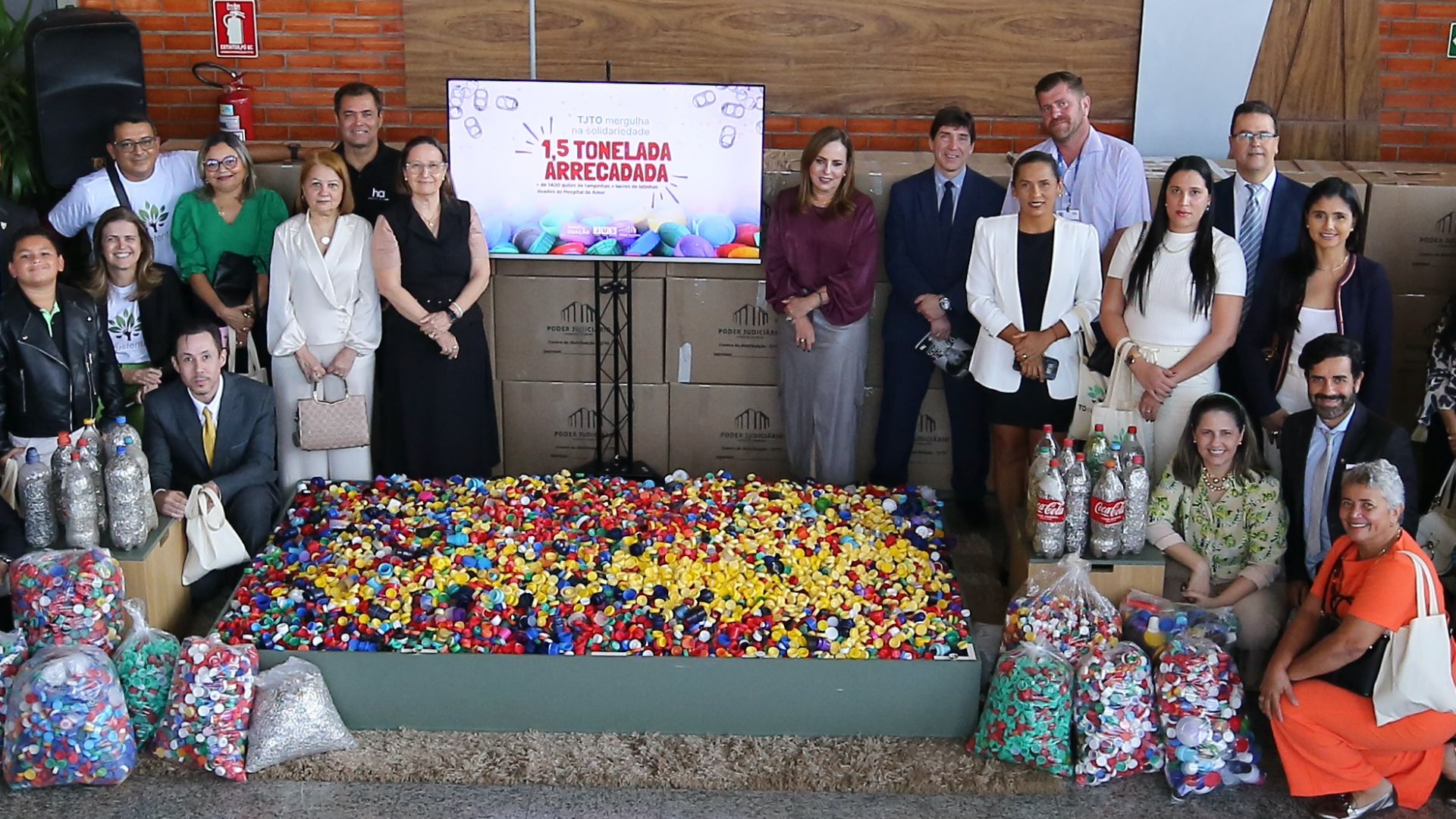Na imagem, vemos um grande grupo de pessoas reunido em um saguão espaçoso com paredes de tijolos aparentes, provavelmente de uma instituição pública ou corporativa, celebrando uma ação solidária. Ao centro, uma tela exibe a frase "1,5 tonelada arrecadada", indicando o sucesso de uma campanha, possivelmente de arrecadação de tampinhas plásticas, como sugerem os vários sacos e a grande quantidade dessas tampinhas coloridas organizadas no chão à frente do grupo. Os participantes, vestindo trajes formais ou sociais, posam sorridentes para a foto coletiva, muitos segurando sacolas ecológicas com o logotipo do evento. O ambiente é moderno, bem iluminado e decorado com plantas, banners comemorativos e retratos na parede, sugerindo também uma celebração pelos 35 anos da instituição anfitriã. O clima geral é de orgulho e comemoração por uma ação comunitária bem-sucedida.