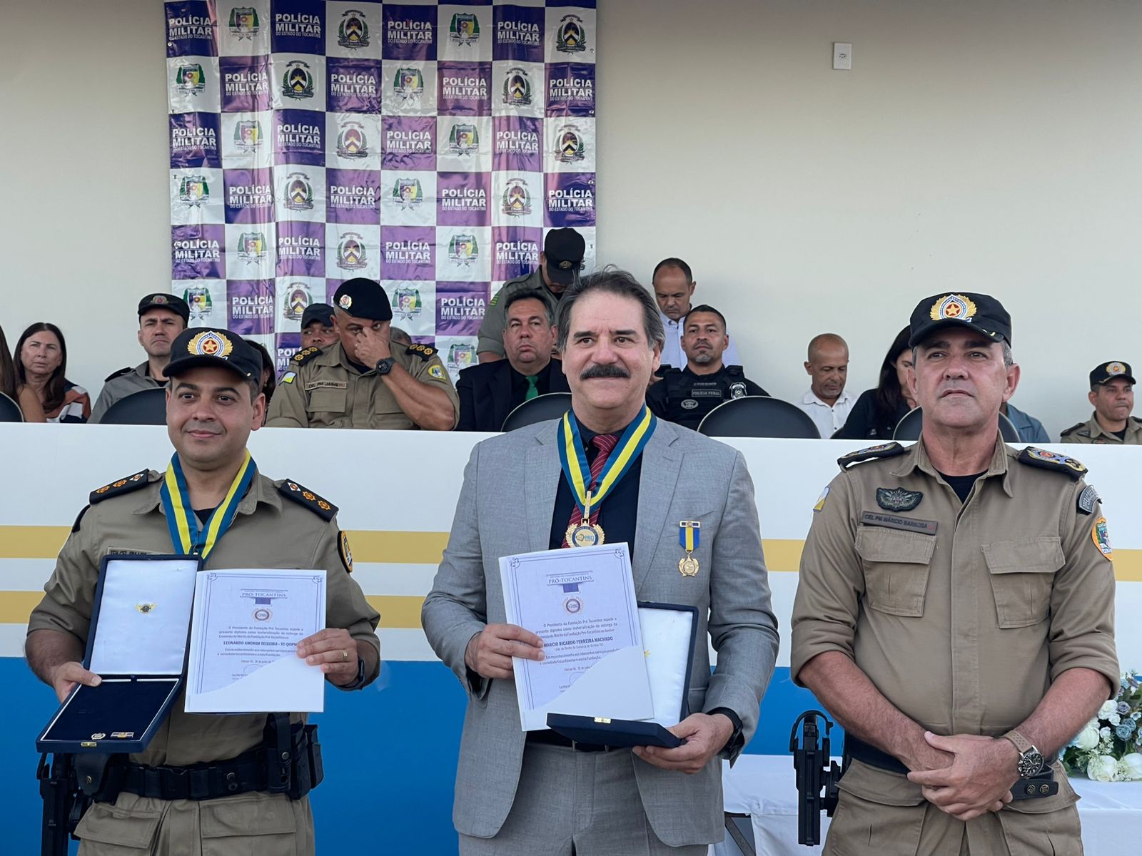 Fotografia colorida onde dois policiais estão um de cada lado, ao lado de um homem que segura um certificado. Ele está usando uma medalha e de terno cinza, com cabelos pretos meio grisalhos e um bigode escuro. Ao fundo, pessoas estão sentadas em um local mais alto. 