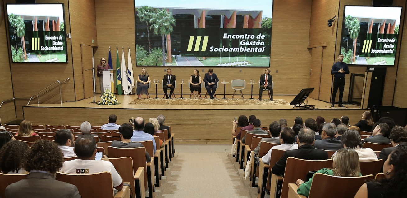 Fotografia colorida de um palco todo composto por madeira clara. Três telas projetam um banner com a foto do Tribunal de Justiça do Tocantins e o nome do evento sobreposto. Em cima do palco, sete pessoas guiam a cerimônia enquanto uma plateia sentada em cadeiras a frente assiste