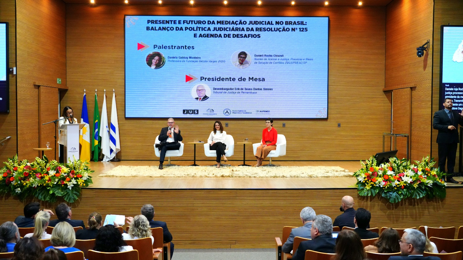 A imagem mostra uma palestra em um auditório, com uma mulher em pé no púlpito falando para uma plateia sentada de costas. Ao fundo, um grande telão exibe o título "O presente e futuro da mediação e conciliação judiciais no Brasil", junto com o logo da FGV Direito SP. À direita do púlpito, dois palestrantes estão sentados em poltronas brancas, um homem de terno e uma mulher de vestido vermelho. Há arranjos florais em ambos os lados do palco.