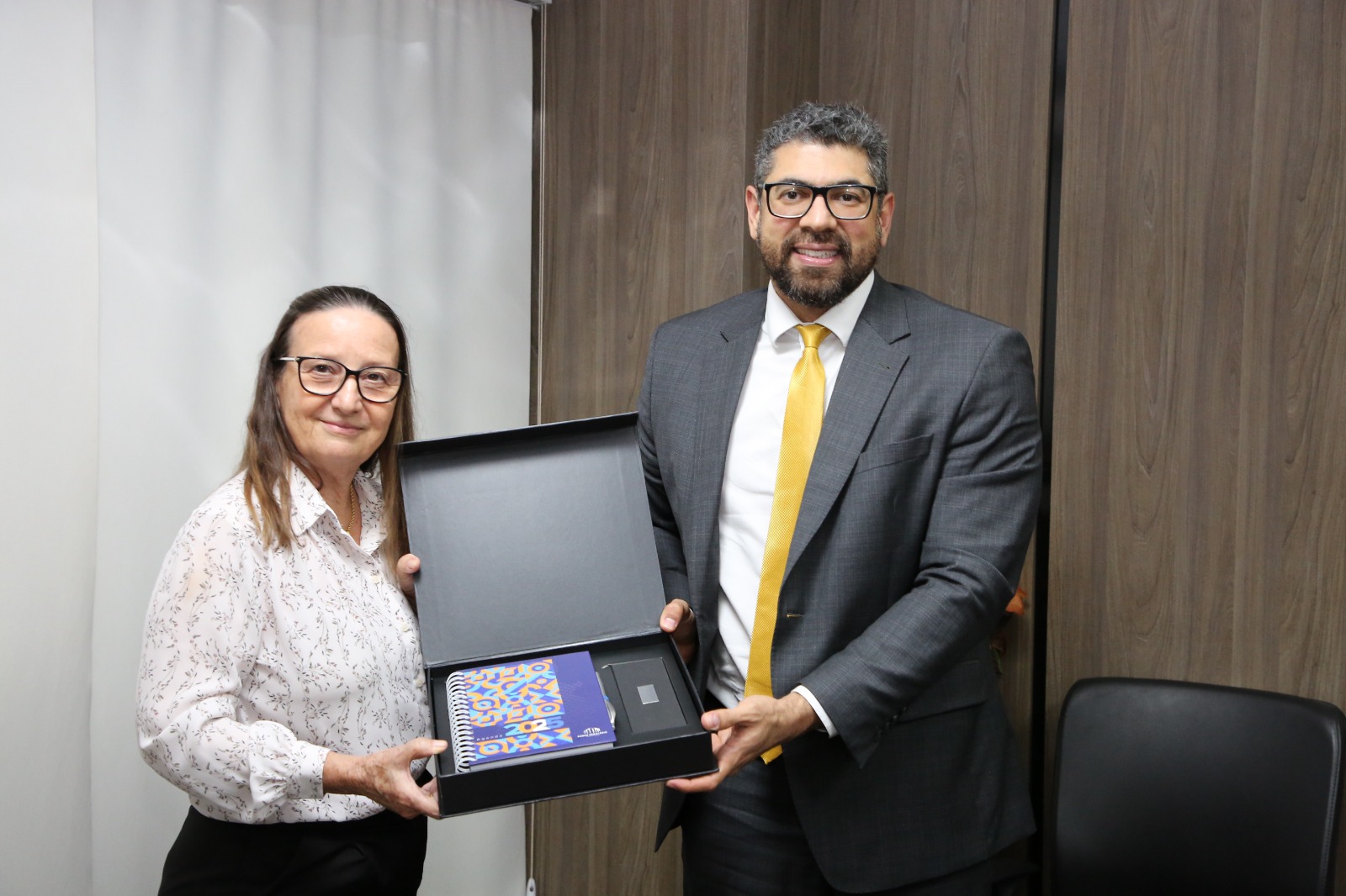 A imagem mostra a desembargadora Maysa entregando uma caixa escura para o secretário Bruno. Ela está à esquerda e ele à direita. Ambos mantêm uma postura formal e atenta. 