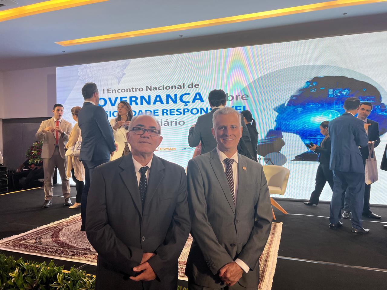 Fotografia colorida onde dois homens estão de pé em frente a um painel branco com escritas em azul. Em sua volta, pessoas conversam. 