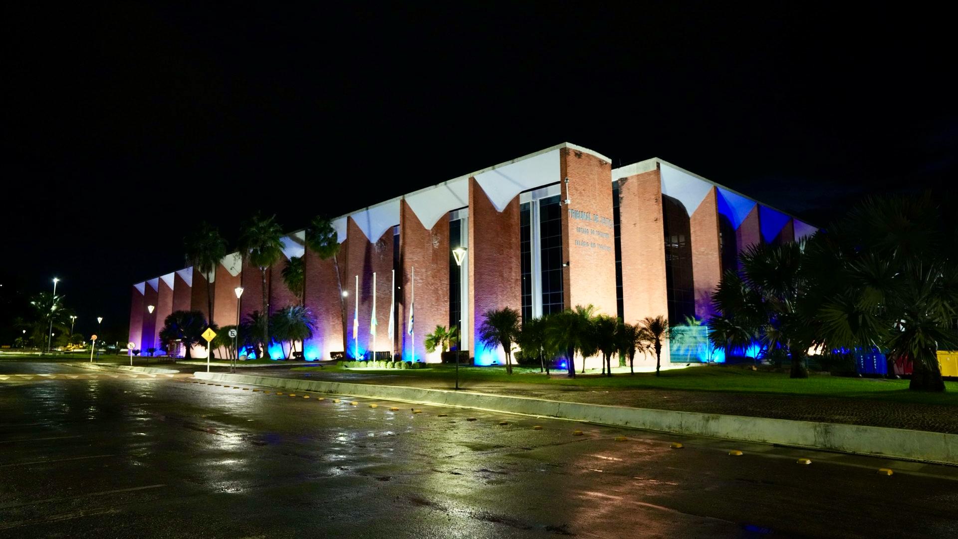 Fotografia colorida do prédio do Tribunal de Justiça do Tocantins de frente. O prédio está iluminado com luzes brancas e azuis, e está de noite. Tudo em volta está escuro. 
