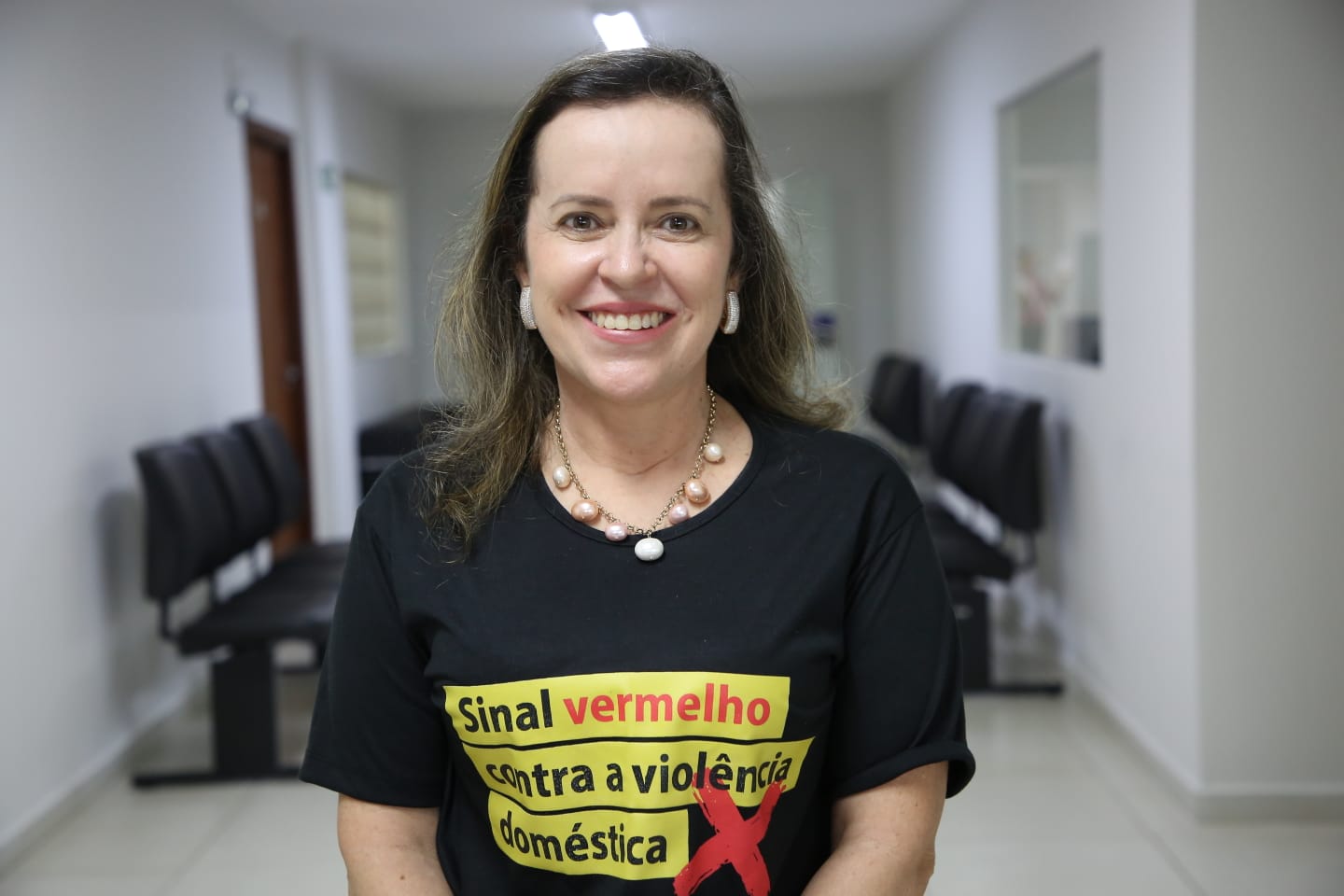 Imagem com uma mulher no centro do enquadramento, e o foco principal está nela. A mulher está posicionada de frente para a câmera. Ela tem cabelos castanhos e um sorriso amigável. O fundo apresenta uma sala com cadeiras de espera em tom de preto, e as paredes são brancas. Há um espaço vazio à direita e à esquerda da mulher.