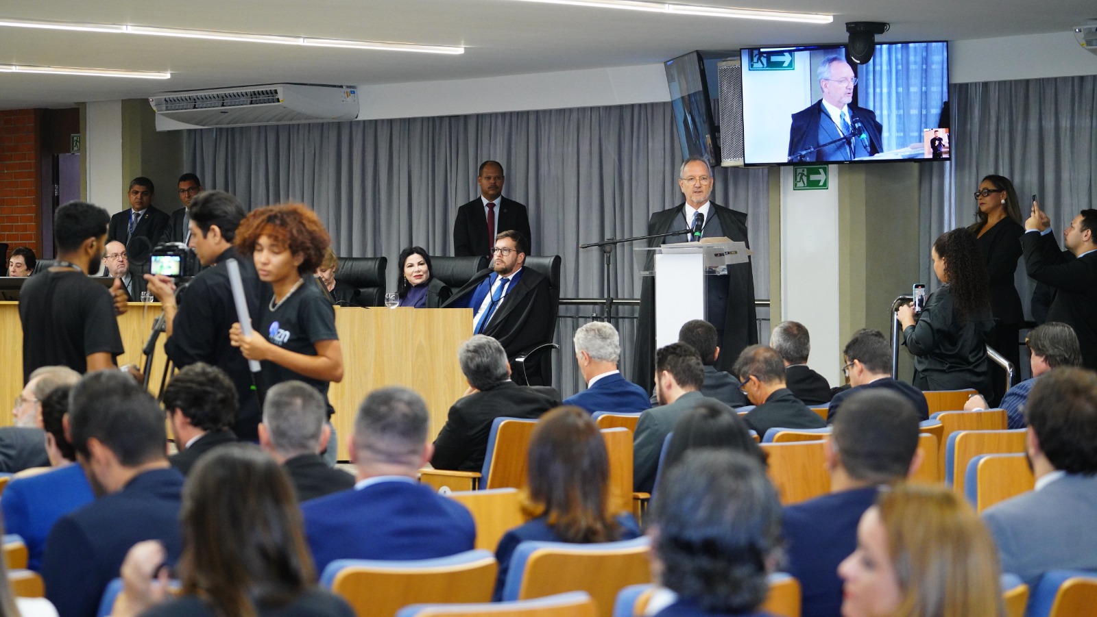 Sessão solene em plenário judicial, com o corregedor-geral, desembargador Pedro Nelson, discursando ao púlpito, magistrados sentados à mesa diretiva, público presente nas cadeiras do auditório e profissionais de imprensa realizando registros audiovisuais. Ambiente interno institucional, com telão exibindo a fala do orador.