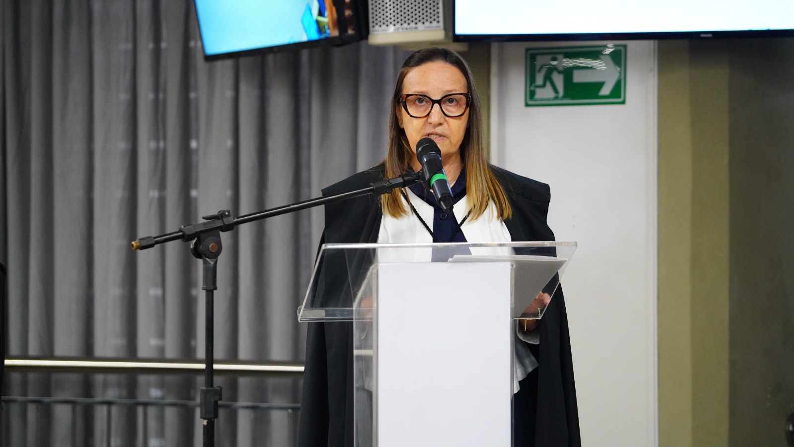 Presidente do TJTO, desembargadora Maysa em trajes de magistratura discursa ao microfone, posicionada atrás de um púlpito transparente, em ambiente interno institucional, com cortina ao fundo e sinalização de saída na parede.