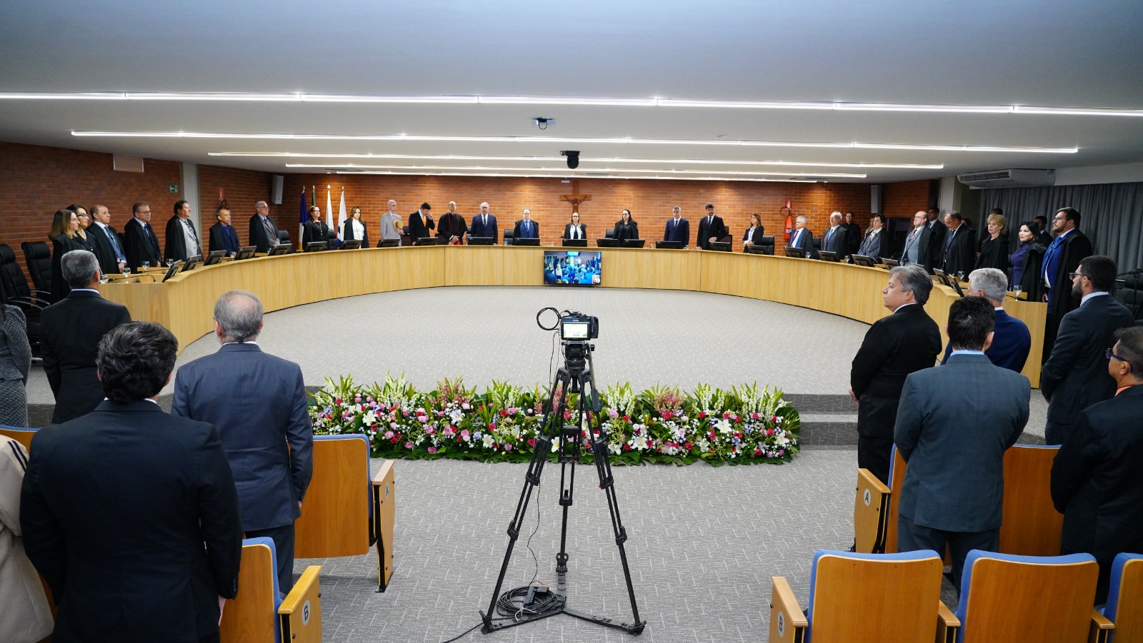 Sessão solene em plenário judicial, com magistrados e autoridades de pé ao redor de mesa semicircular, câmera em tripé no centro do espaço e arranjo floral à frente. Ambiente interno amplo, com iluminação no teto e público presente.