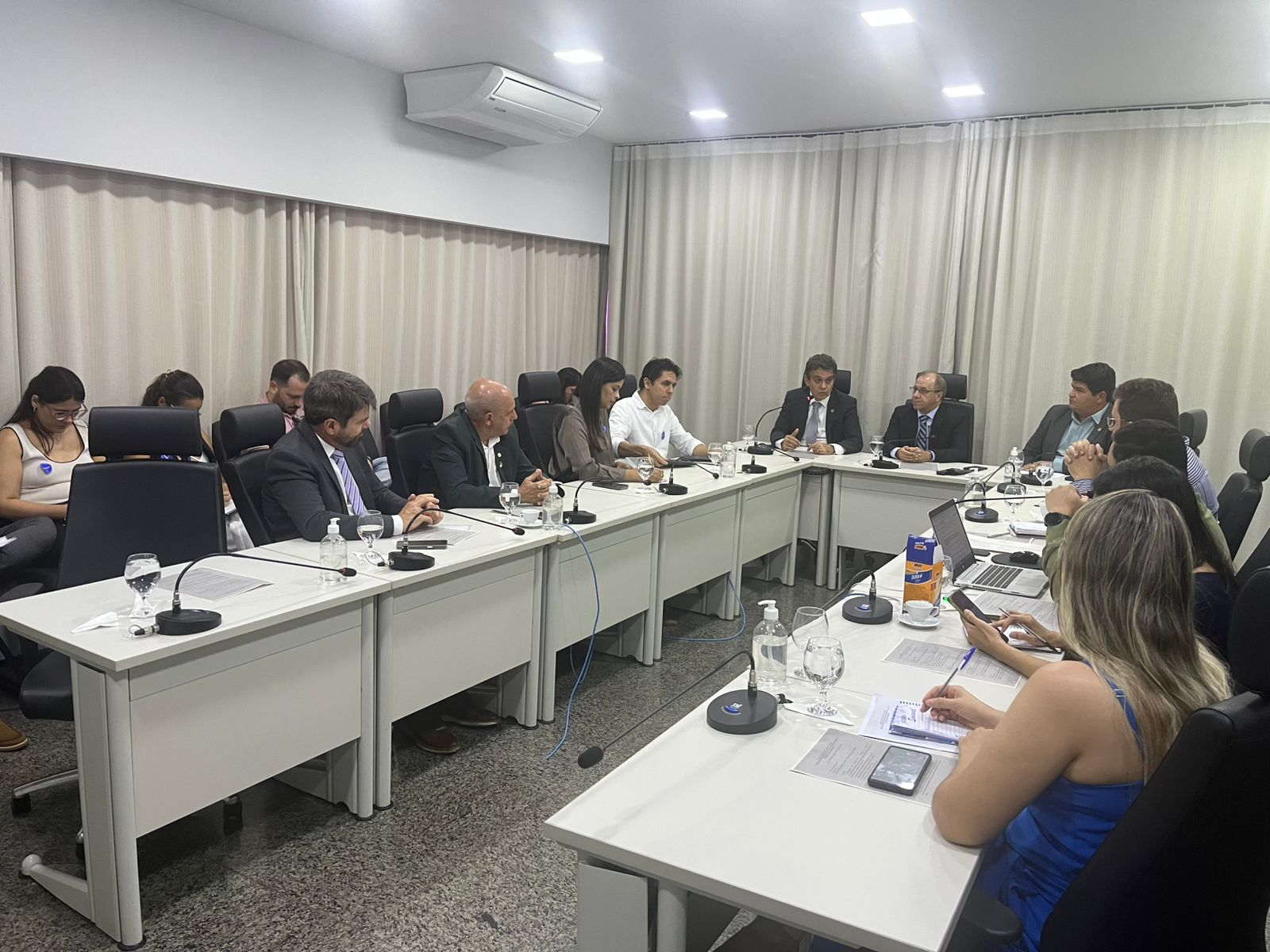 Reunião institucional realizada em sala de reuniões do Tribunal de Justiça do Tocantins, com participantes sentados ao redor de uma mesa em formato de U. Os presentes utilizam trajes formais e acompanham a discussão com documentos, blocos de anotações, notebooks e celulares sobre a mesa. O ambiente é fechado, com iluminação artificial, cortinas claras ao fundo, aparelhos de ar-condicionado e microfones individuais distribuídos ao longo da mesa. Há copos com água, garrafas e materiais de apoio.