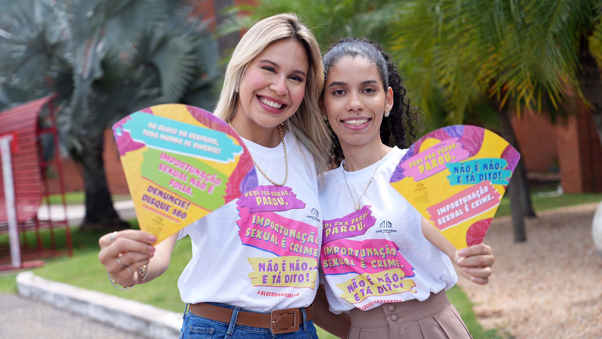 Duas mulheres posam lado a lado, sorrindo para a câmera, em um ambiente externo com árvores e prédio ao fundo. Elas vestem camisetas brancas com identidade visual colorida de campanha educativa, trazendo frases como “Importunação sexual é crime” e “Não é não. E tá dito!”.  Cada uma segura um leque personalizado, também em cores vibrantes, com mensagens de conscientização contra a importunação sexual e incentivo à denúncia, incluindo o número “Disque 180”. A cena transmite um clima leve e acolhedor, reforçando a mensagem de respeito e combate à violência durante eventos festivos.