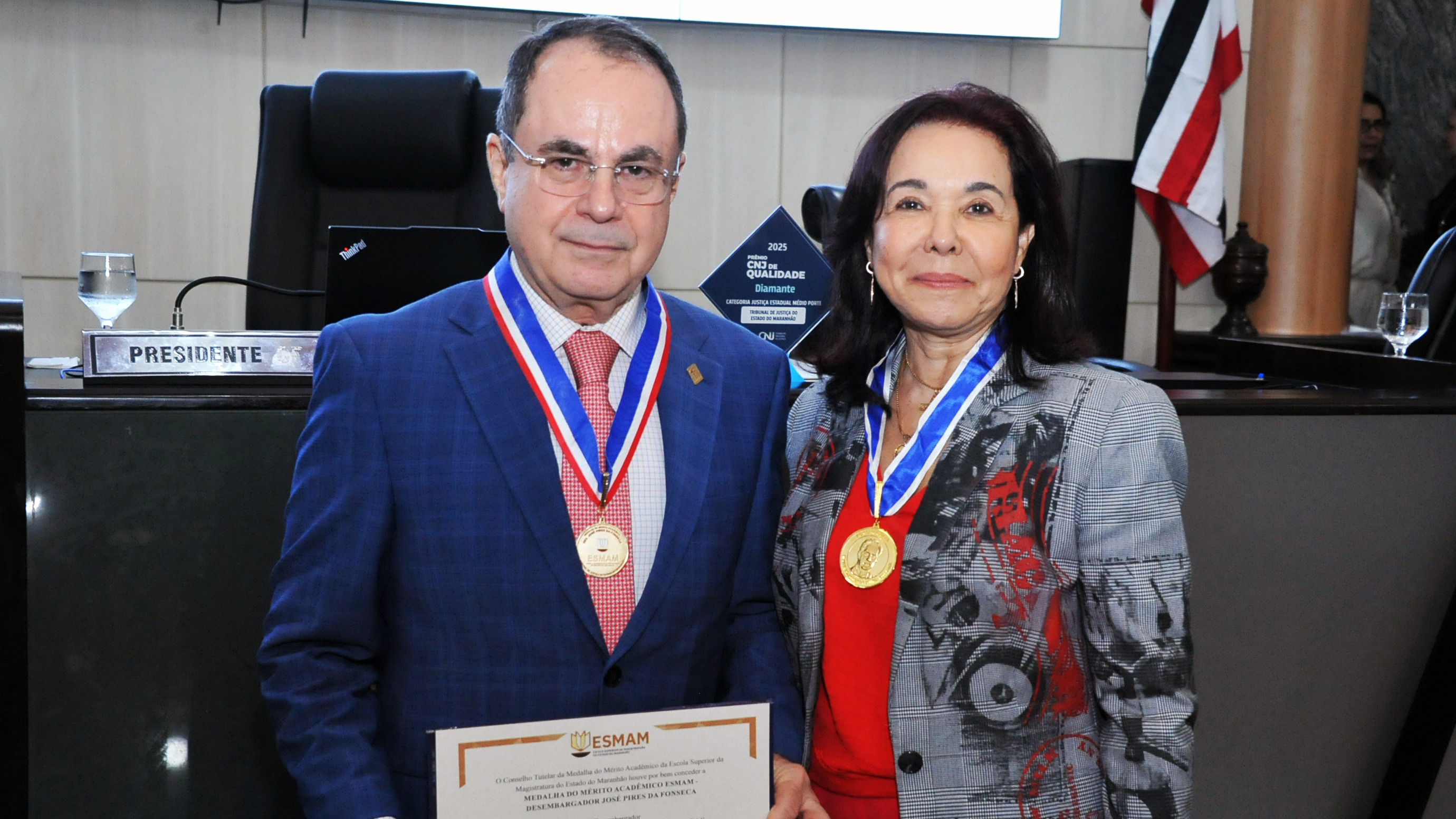 A imagem mostra dois magistrados, um homem e uma mulher, em cerimônia oficial no plenário do Judiciário, usando medalhas de condecoração. O homem segura um certificado da ESMAM. O ambiente é solene, com mesa diretiva e identificação de “Presidente” ao fundo, indicando evento institucional de reconhecimento.