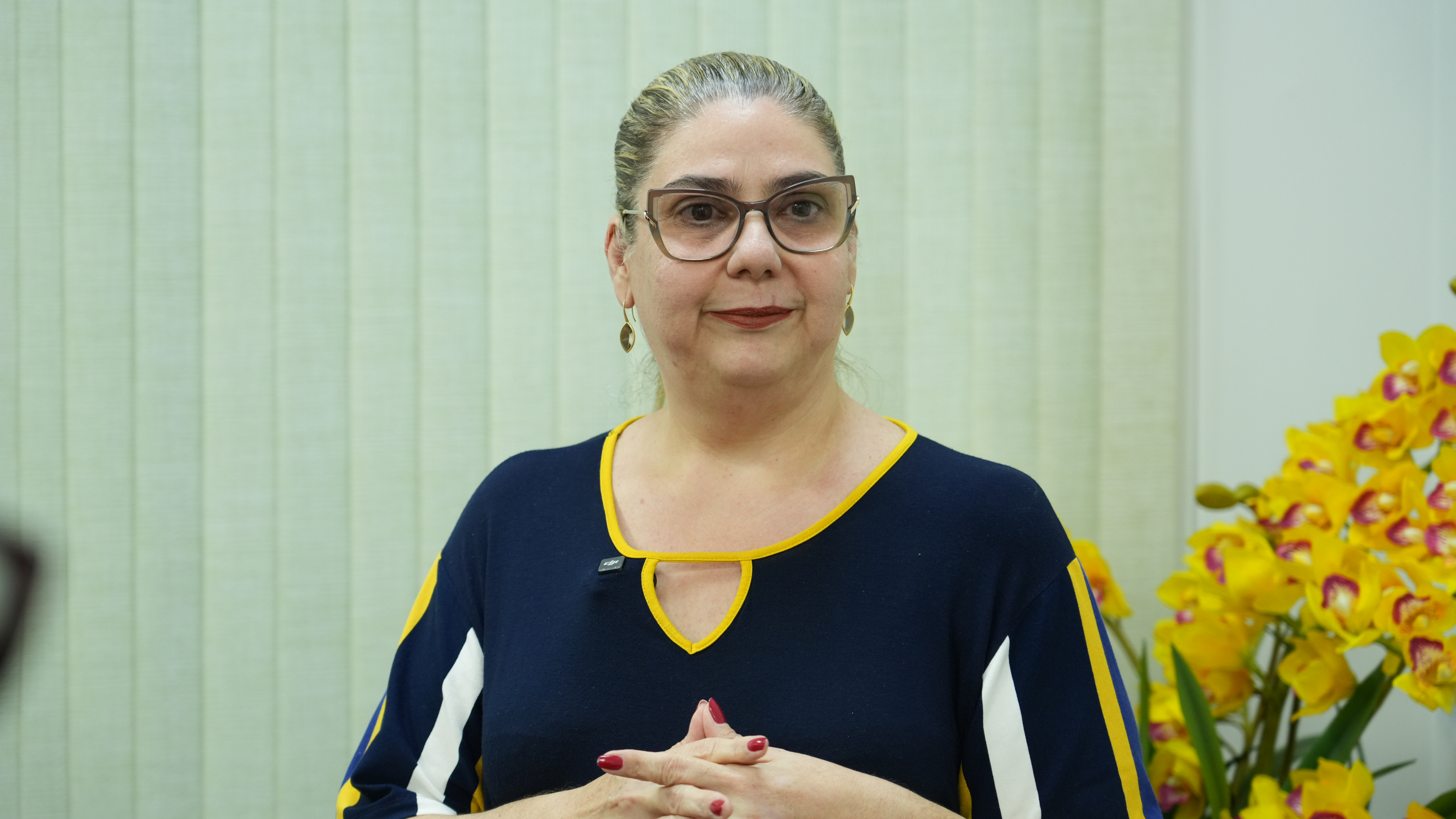 Juíza Flávia Bovo em ambiente interno, posicionada diante de persianas verticais claras. Ela usa óculos, brincos dourados e blusa azul-marinho com detalhes em amarelo e branco. Está com as mãos unidas à frente do corpo. Ao lado direito da imagem, há um arranjo de flores amarelas.
