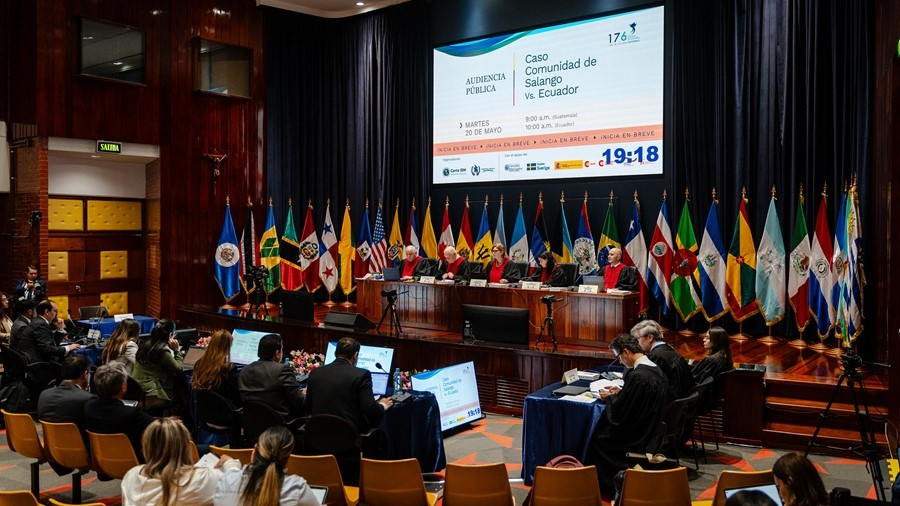 A imagem mostra autoridades reunidas na Corte Interamericana de Direitos Humanos, sentadas em uma bancada, com diversas bandeiras de países ao fundo. À frente, participantes acompanham a sessão enquanto um telão exibe informações sobre o caso Comunidade de Salango vs. Equador.
