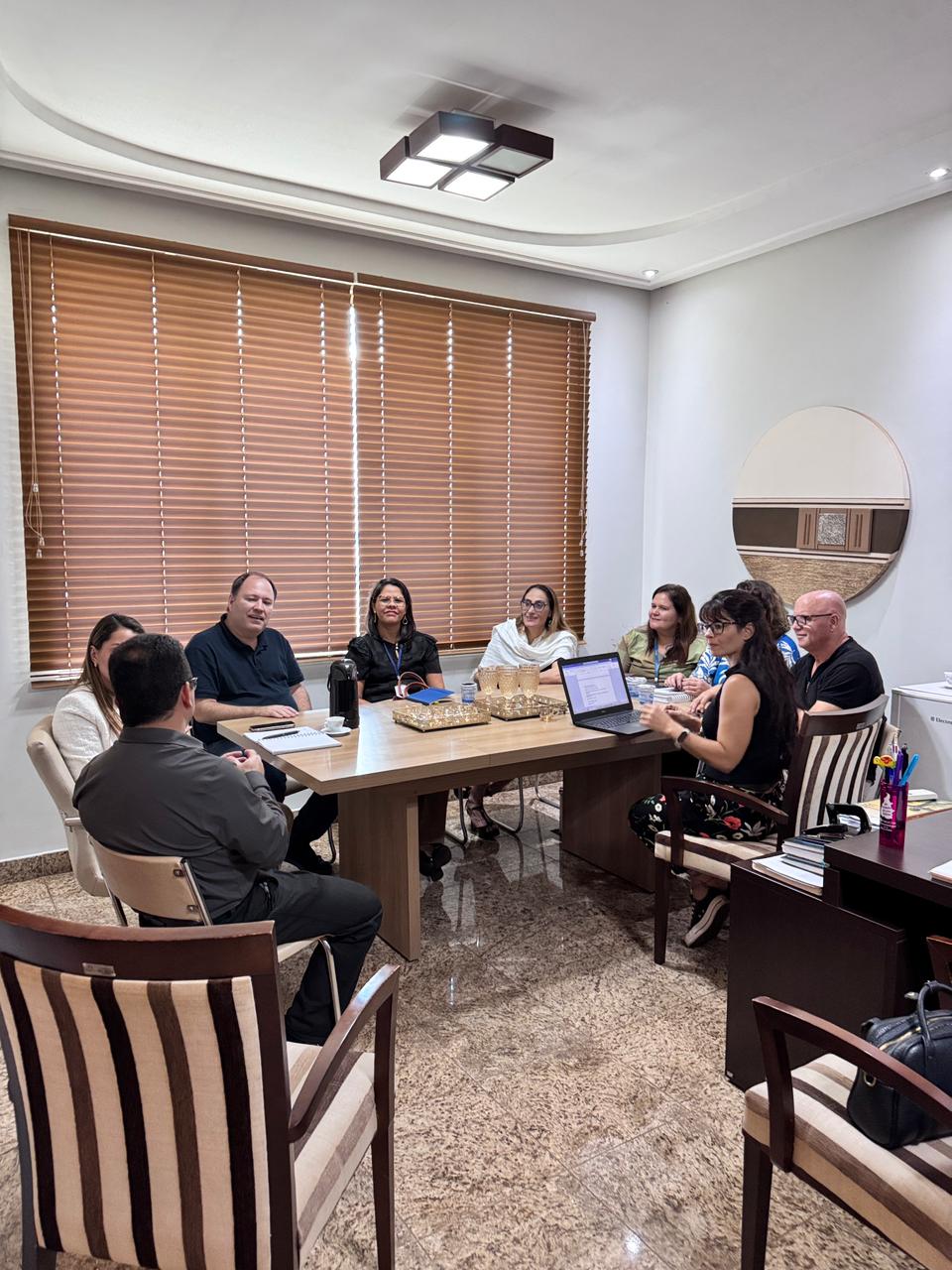 Representantes de instituições participam de uma reunião em uma sala de trabalho, sentados ao redor de uma mesa retangular de madeira. O grupo conversa enquanto analisa informações apresentadas em um notebook aberto sobre a mesa. O ambiente é iluminado e organizado, com persianas de madeira cobrindo a janela ao fundo e objetos de apoio, como cadernos, garrafas térmicas e bandejas, distribuídos sobre a mesa. A cena retrata um momento de diálogo e planejamento entre os participantes.