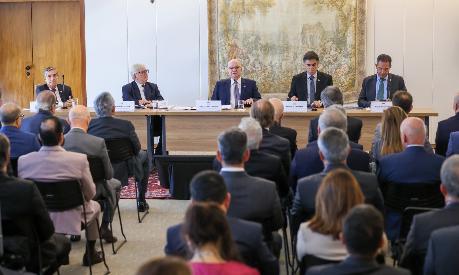 Ministros do Supremo Tribunal Federal participam de mesa de debate em evento institucional, diante de plateia composta por autoridades e convidados. No centro da mesa está o ministro Luiz Edson Fachin, com identificação em placa, ao lado de outros ministros. O encontro ocorre em auditório, com público acomodado em fileiras e painel artístico decorativo ao fundo.