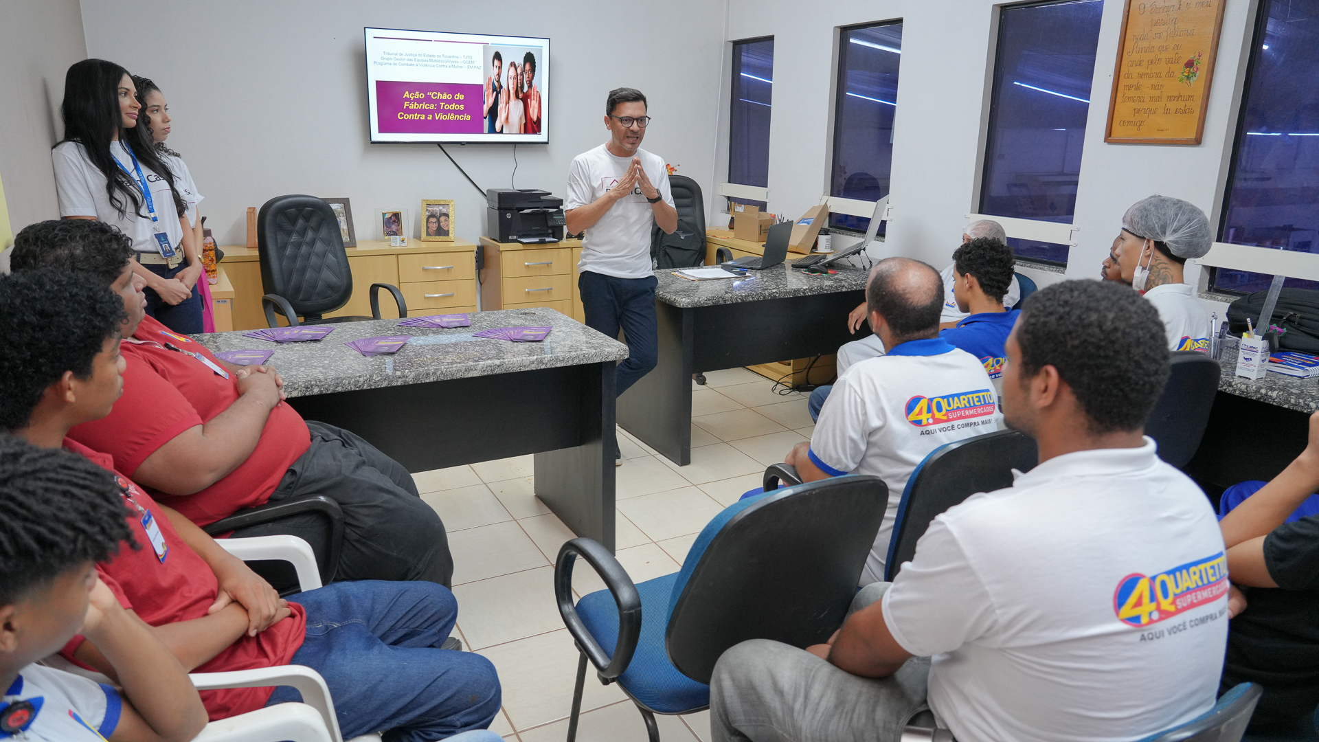 A imagem mostra uma roda de conversa realizada em um ambiente de trabalho, provavelmente em uma sala administrativa de um supermercado. Um homem em pé, ao centro, conduz a conversa com um grupo de funcionários que estão sentados em cadeiras ao redor de duas mesas. Ele gesticula com as mãos enquanto fala, indicando que está explicando ou orientando os participantes.  Ao fundo, um monitor exibe uma apresentação com o tema “Ação Chão de Fábrica: Todos Contra a Violência”, sugerindo que o encontro trata de conscientização ou prevenção à violência. Sobre as mesas há alguns folhetos distribuídos aos participantes.  Os trabalhadores vestem uniformes do supermercado, e alguns utilizam equipamentos como touca e máscara, o que indica que atuam em setores de manipulação de alimentos. À esquerda, duas mulheres acompanham a atividade em pé.