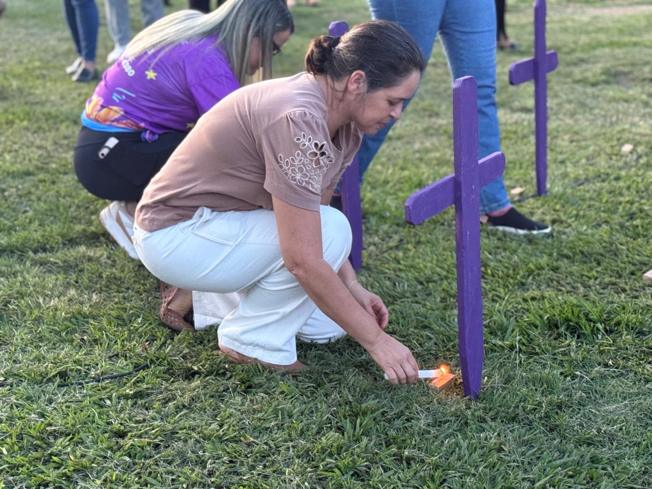 Uma mulher agachada acende uma vela ao lado de uma cruz roxa fincada na grama durante o ato de memória pelas vítimas de feminicídio realizado no Parque Mutuca, em Gurupi. Ao fundo, outra participante também se ajoelha diante de uma das cruzes, em um momento de silêncio e reflexão que marcou a homenagem às mulheres vítimas de violência.
