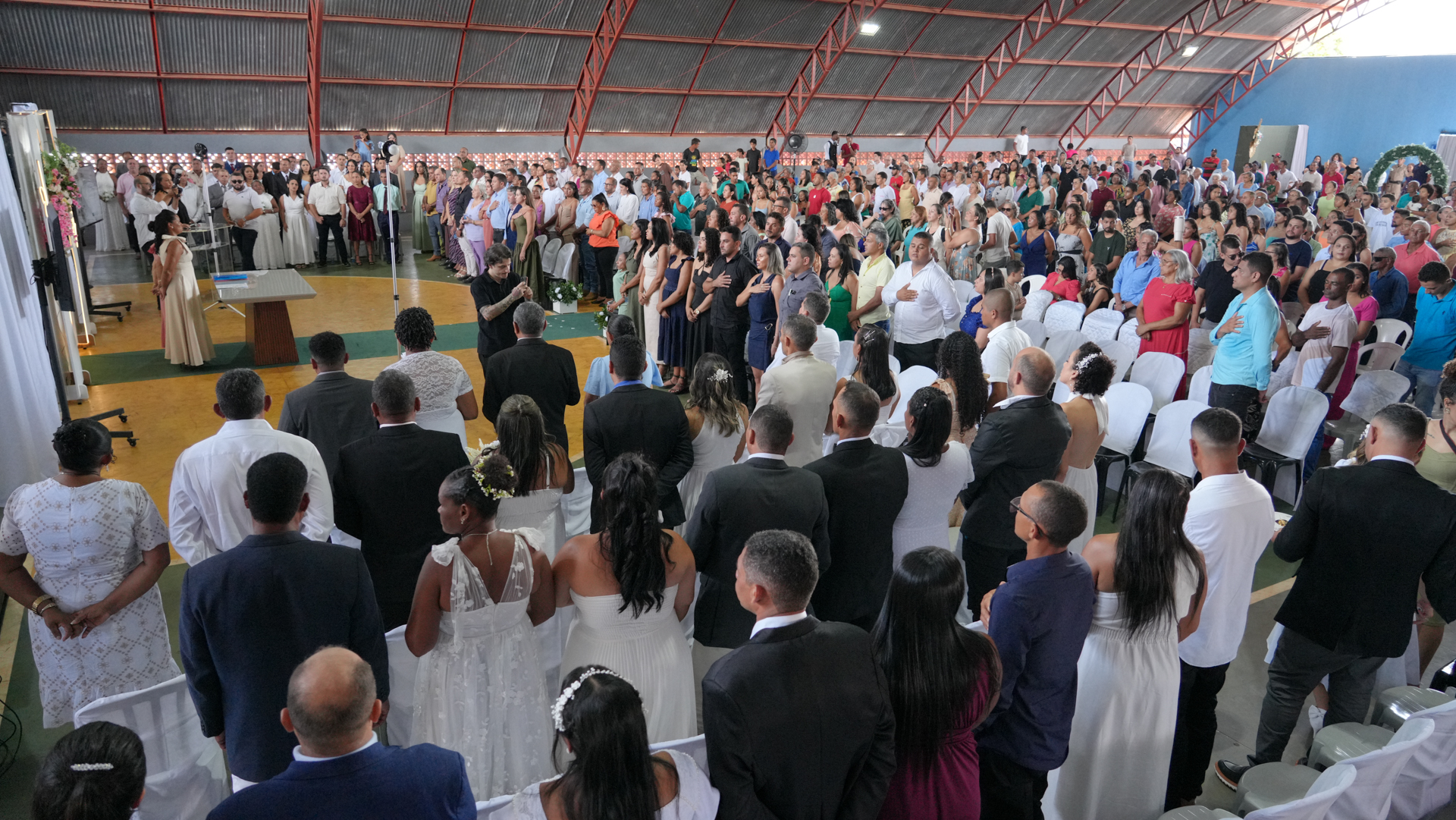 Imagem ampla de um casamento comunitário em um ginásio poliesportivo, com dezenas de casais diante do altar e grande presença de familiares e convidados acompanhando a cerimônia