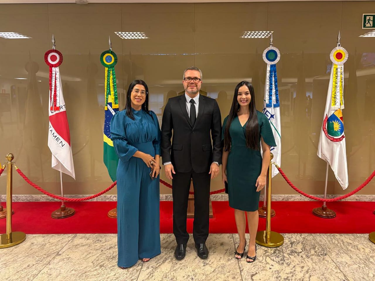 O juiz Marcelo Laurito posa para foto ao lado das servidoras Dayse Santos Maciel e Sabrina Assakawa Ludgero Leal em espaço institucional. Ao fundo, aparecem bandeiras oficiais, incluindo a do Brasil, em ambiente interno com piso de mármore e cordões de isolamento.