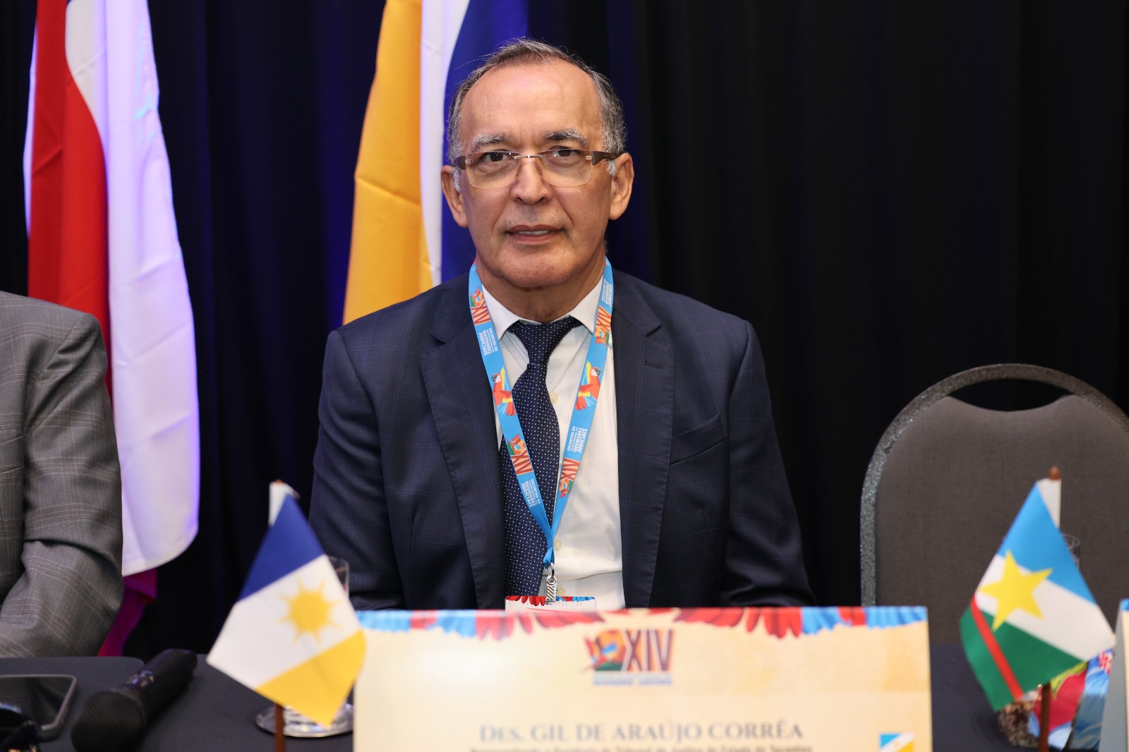 Desembargador Gil Correa sentado à mesa de evento, com identificação nominal à frente, vestindo terno escuro e gravata. Usa crachá e está posicionado entre bandeiras, em ambiente institucional.