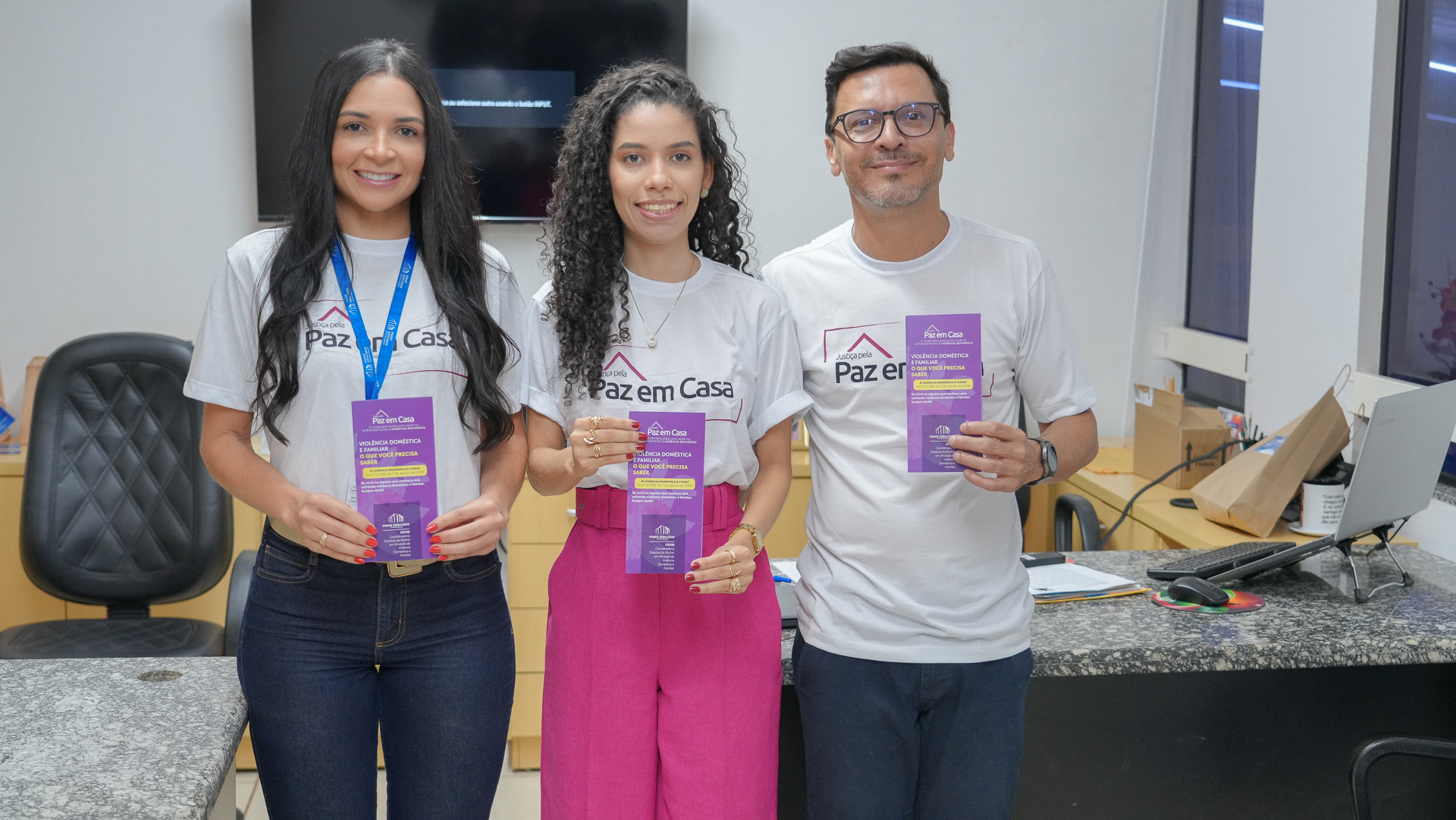 Três pessoas estão em ambiente interno de trabalho, lado a lado, sorrindo e olhando para a câmera. Todas vestem camisetas da campanha “Paz em Casa” e seguram materiais informativos sobre violência doméstica. Ao fundo, há uma mesa de escritório com computador, cadeiras e itens de trabalho, indicando um espaço institucional