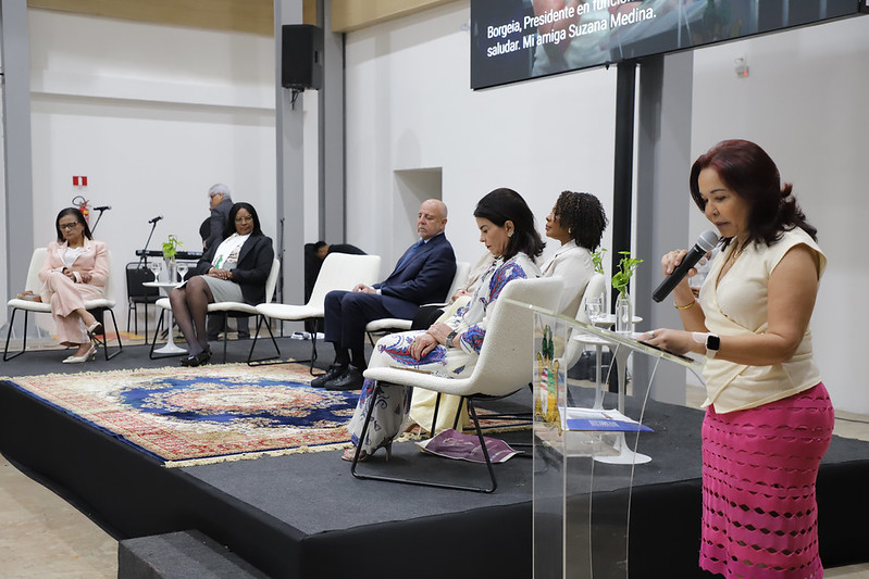 A imagem mostra o dispositivo de honra na abertura da Conferência Internacional “Mulheres Juízas em Conexão”, com autoridades sentadas no palco, enquanto uma mulher faz uso da palavra em um púlpito.