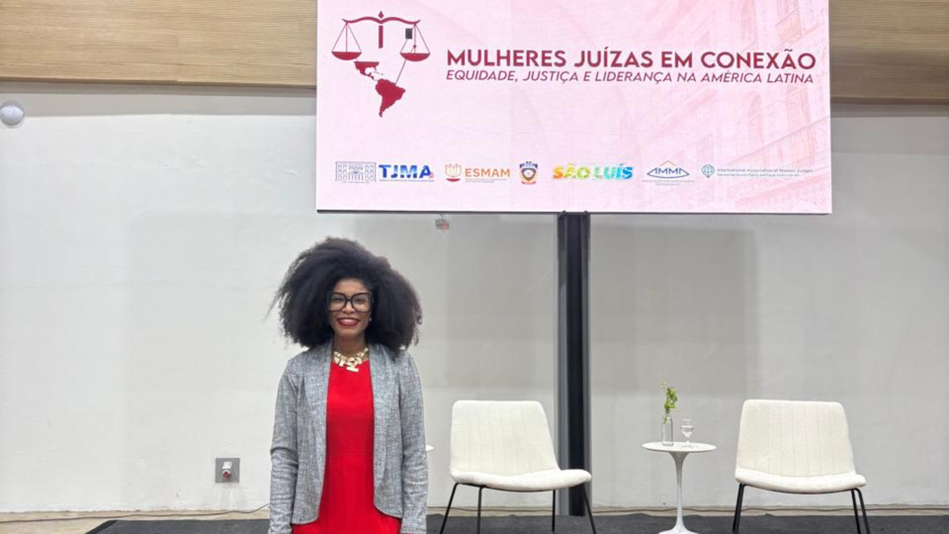A imagem mostra a juíza Renata do Nascimento durante a Conferência Internacional “Mulheres Juízas em Conexão”. A magistrada aparece com cabelo solto, usando vestido vermelho, blazer cinza e sapatos pretos, posando em frente ao painel do evento.