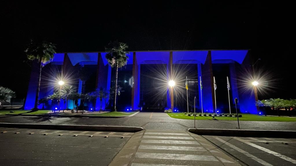 Prédio-sede do Tribunal de Justiça do Tocantins iluminado em azul durante a noite. A fachada moderna é destacada por luzes azuis que cobrem as colunas e a entrada principal. À frente do edifício, há um caminho com faixa de pedestres, gramado, palmeiras e mastros com bandeiras. O céu está escuro, evidenciando a iluminação especial da estrutura.