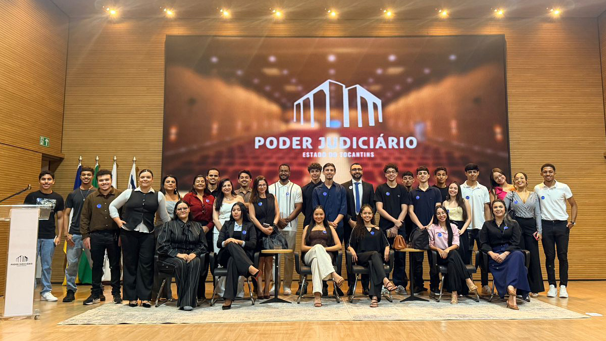 Grupo de estudantes e profissionais posa para foto no auditório do TJTO. Ao fundo, painel com a identidade institucional do Tribunal, indicando visita guiada