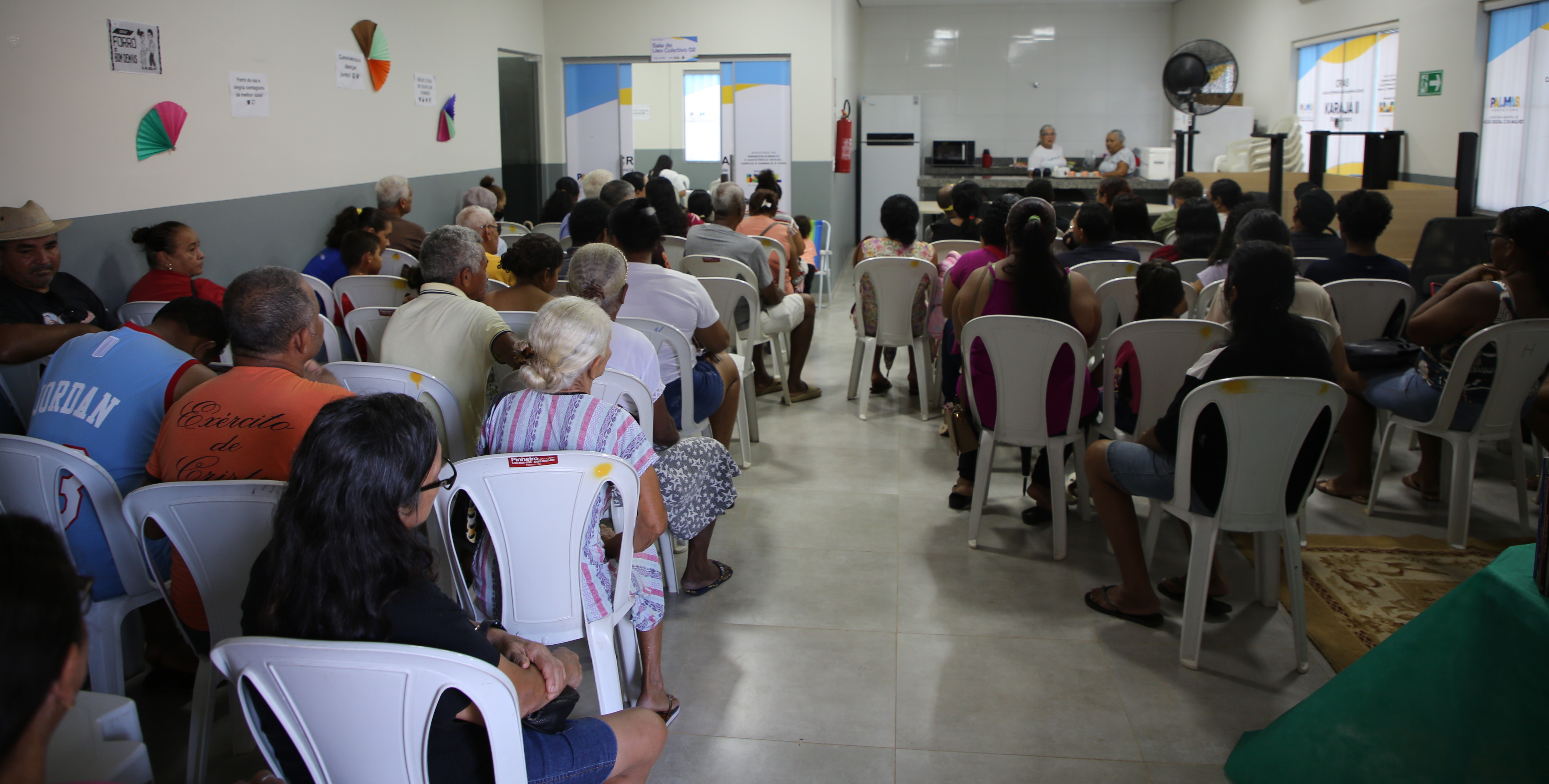 Sala com diversas pessoas sentadas em cadeiras, participando de atendimento ou reunião coletiva. Ao fundo, duas atendentes realizam orientações em um espaço organizado, indicando ação social ou prestação de serviços à comunidade