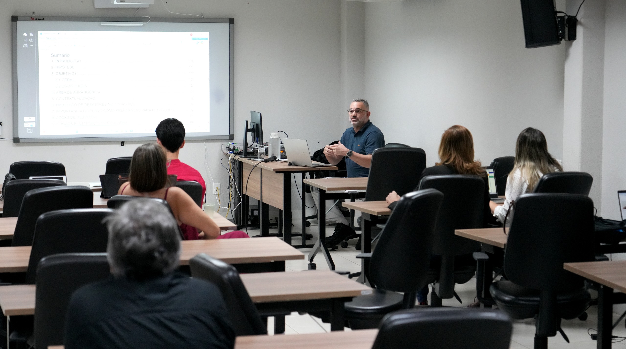 A imagem mostra um instrutor ministrando aula em uma sala, sentado à frente com computador, enquanto participantes acompanham atentos em suas cadeiras. A cena integra o curso de elaboração do Plano de Contingência Socioambiental do Poder Judiciário do Tocantins.