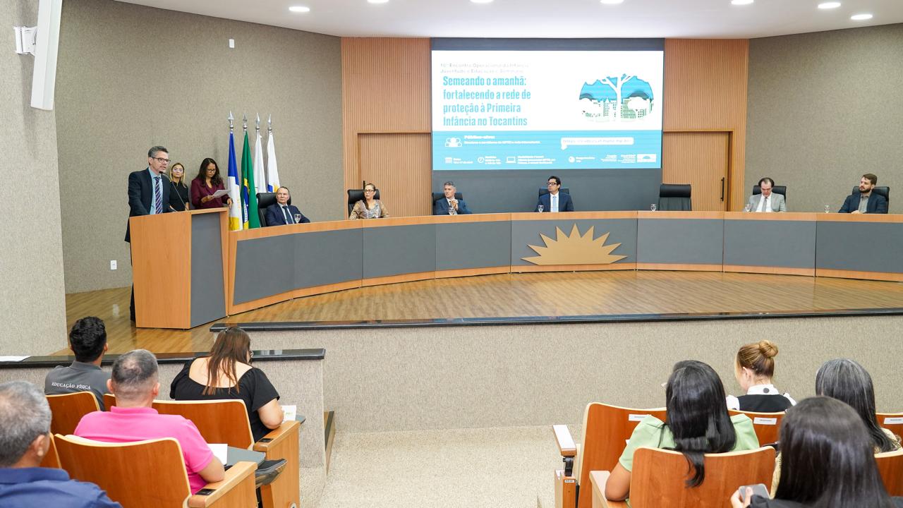 A imagem mostra autoridades do Judiciário e representantes de instituições reunidos em auditório durante seminário sobre o fortalecimento da proteção à primeira infância no Tocantins. No palco, a mesa de honra está composta enquanto o juiz Adriano Gomes de Melo faz uso da palavra ao microfone, com o público atento à programação.