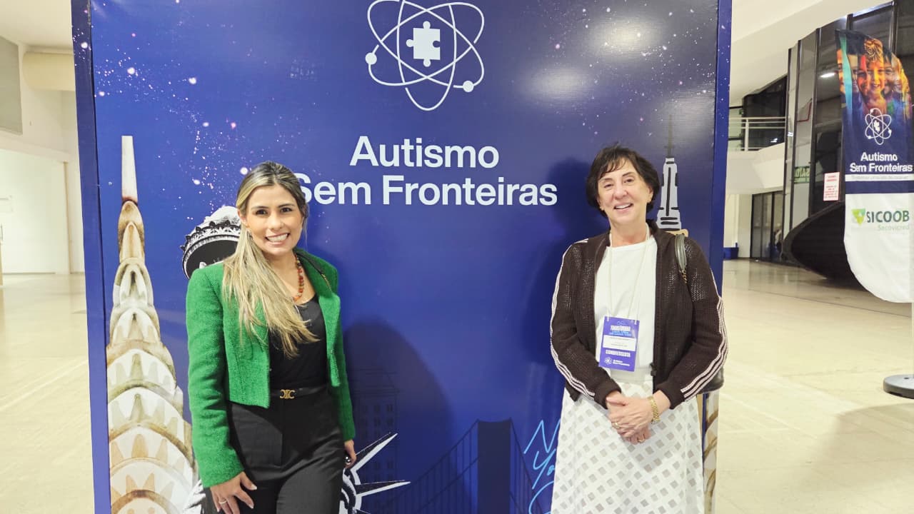 A desembargadora Ângela Haonat e uma servidora do Tribunal de Justiça do Tocantins posam para foto em frente ao painel do evento “Autismo Sem Fronteiras”. Ambas sorriem para a câmera. A magistrada usa crachá de identificação do congresso.