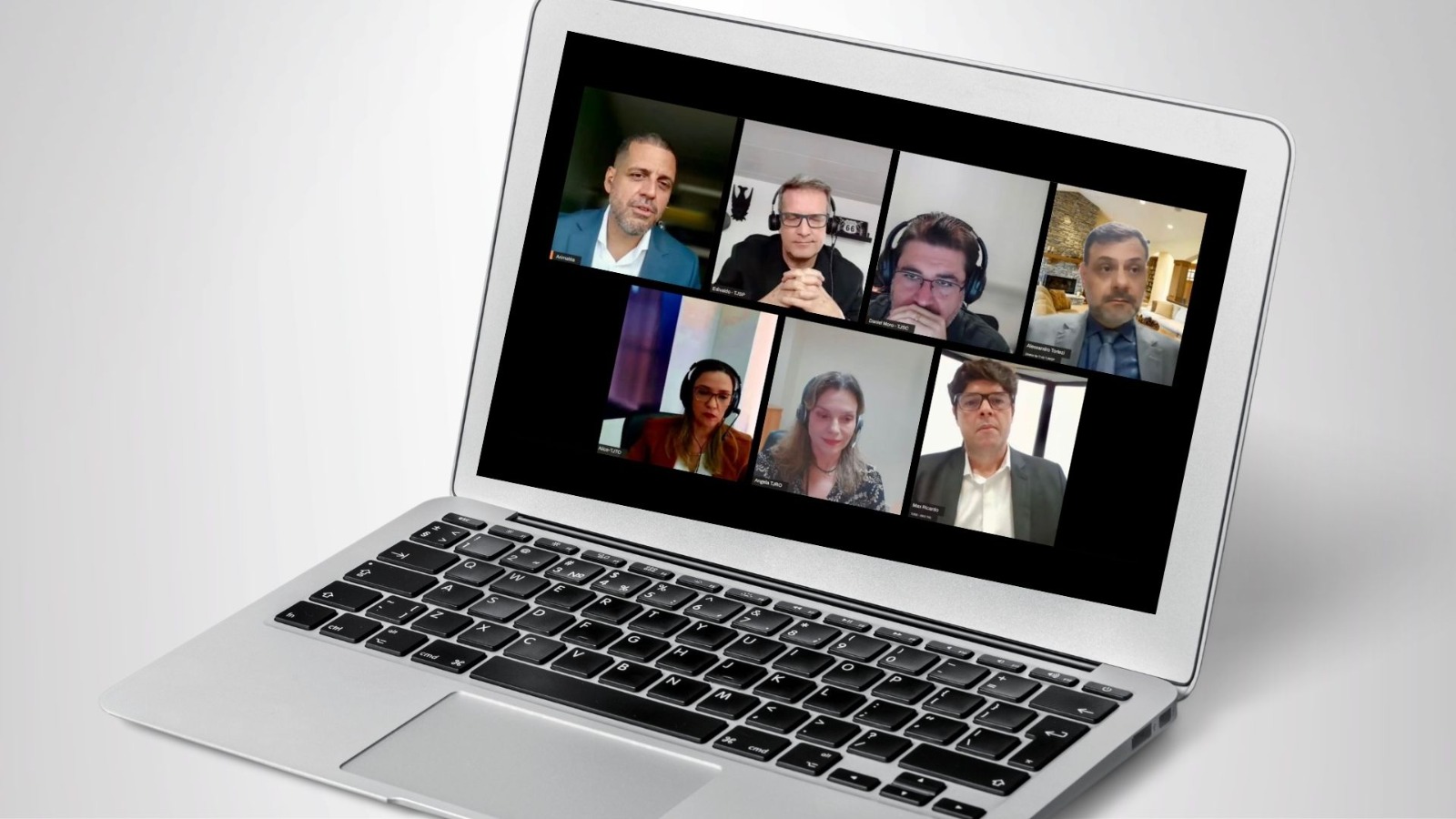 Notebook aberto exibindo uma videoconferência com oito participantes em formato de grade. Os convidados aparecem em suas câmeras, alguns usando fones de ouvido, em ambientes internos variados. A tela sugere um debate online, com enquadramento típico de transmissão ao vivo ou reunião virtual.