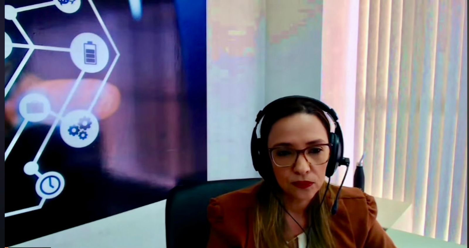 A diretora de Tecnologia da Informação do TJTO, Alice Setúbal, aparece em primeiro plano durante participação em videoconferência. Ela utiliza fones de ouvido com microfone e veste blazer em tom terroso, mantendo postura atenta diante da câmera. Ao fundo, um painel com elementos gráficos que remetem à tecnologia — como ícones de conexão, engrenagens e bateria. À direita, uma janela com persianas verticais ilumina o ambiente, compondo um cenário corporativo e organizado.