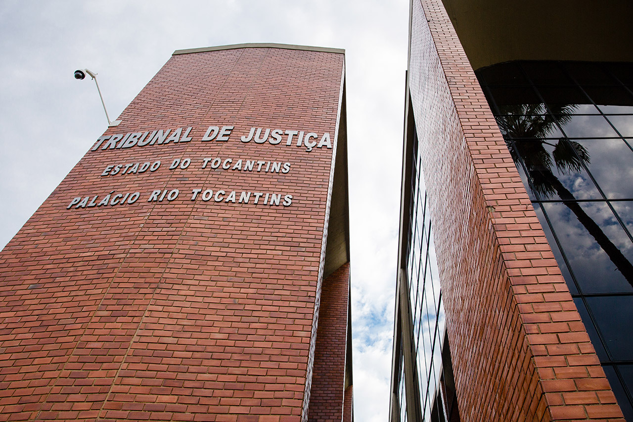Fachada do Tribunal de Justiça do Tocantins, com parede de tijolos aparentes e inscrição “Tribunal de Justiça – Estado do Tocantins – Palácio Rio Tocantins”. À direita, estrutura com vidro refletindo o céu e uma palmeira.