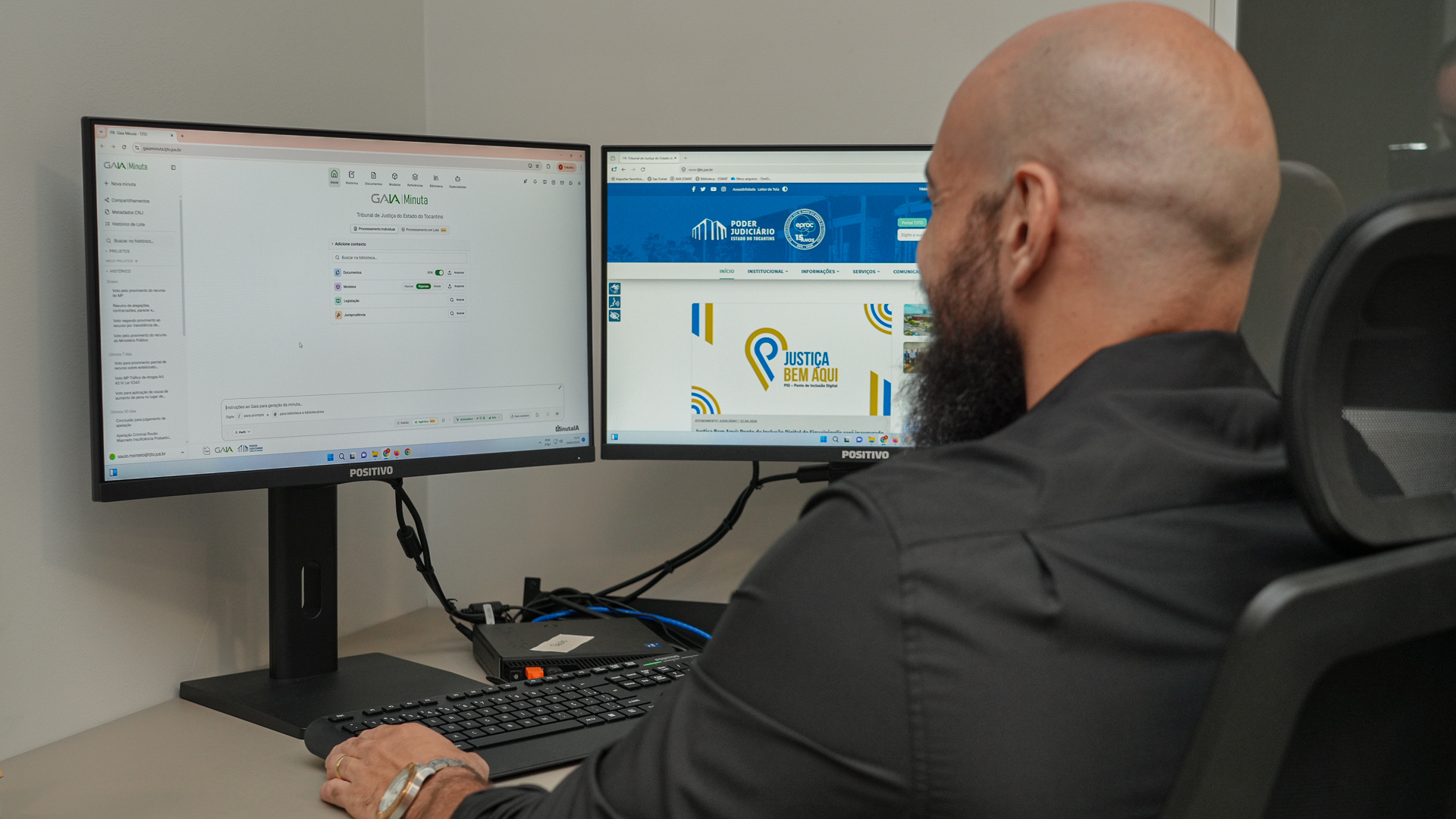 Servidor trabalha em estação com dois monitores. Em um deles, está aberta a interface do sistema GAIA Minuta; no outro, o portal do Poder Judiciário do Tocantins, com destaque para a campanha “Justiça Bem Aqui”. Ele utiliza teclado e mouse diante de um computador desktop, em ambiente de escritório.