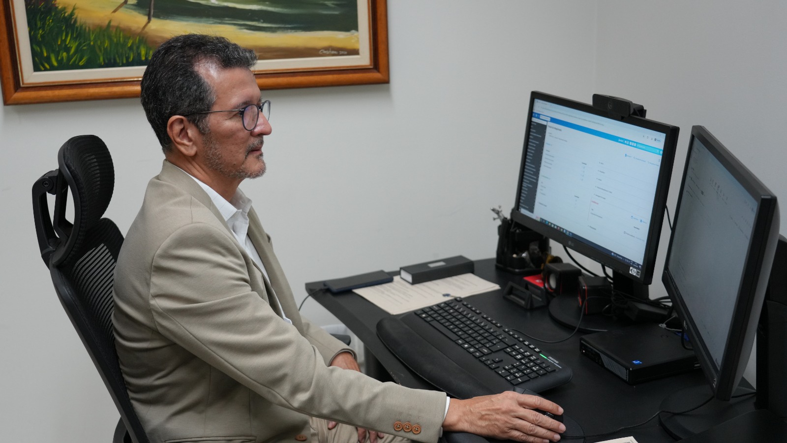 Juiz Rafael trabalha em seu gabinete, diante de uma estação com dois monitores, utilizando teclado e mouse. Ele veste blazer claro e óculos, concentrado na tela que exibe sistema digital de gestão processual.