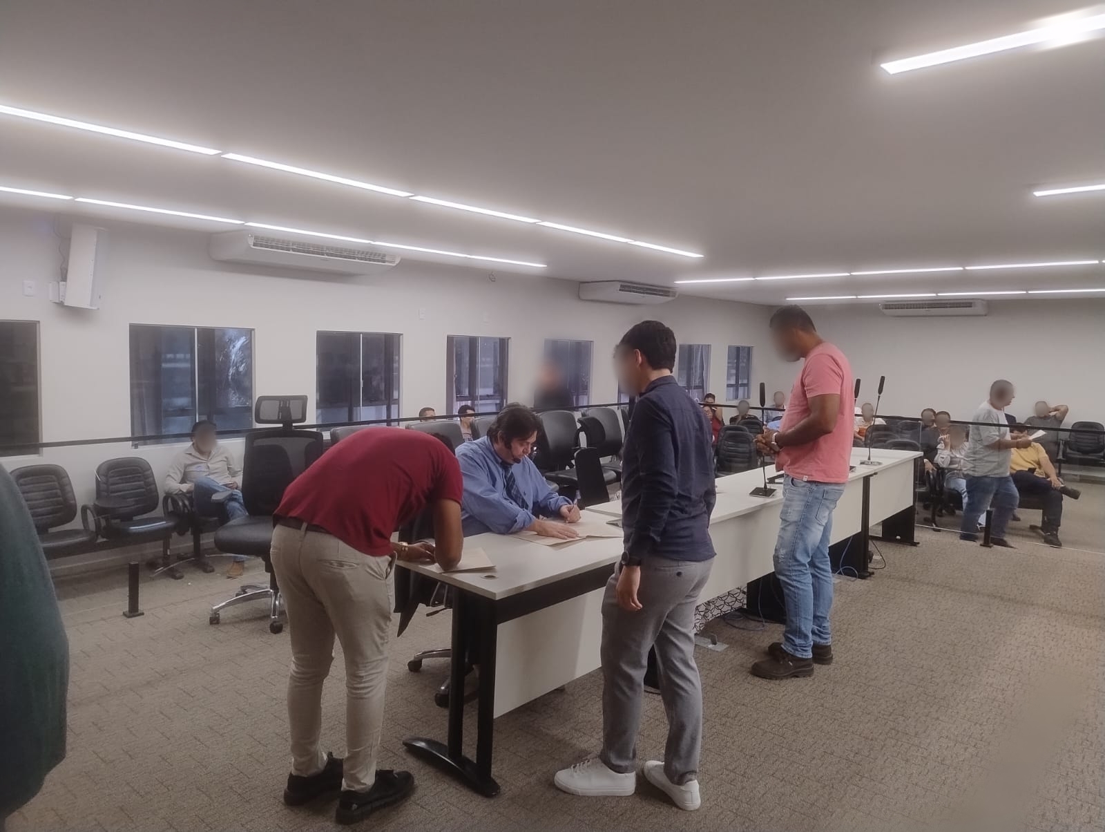 Audiência realizada em sala ampla e iluminada, com o juiz ao centro da mesa, vestido com camisa azul, analisando documentos. À frente dele, duas pessoas assinam papéis, enquanto um terceiro participante aguarda em pé ao lado. Ao fundo, outros presentes acompanham a sessão sentados, em cadeiras organizadas em fileiras.