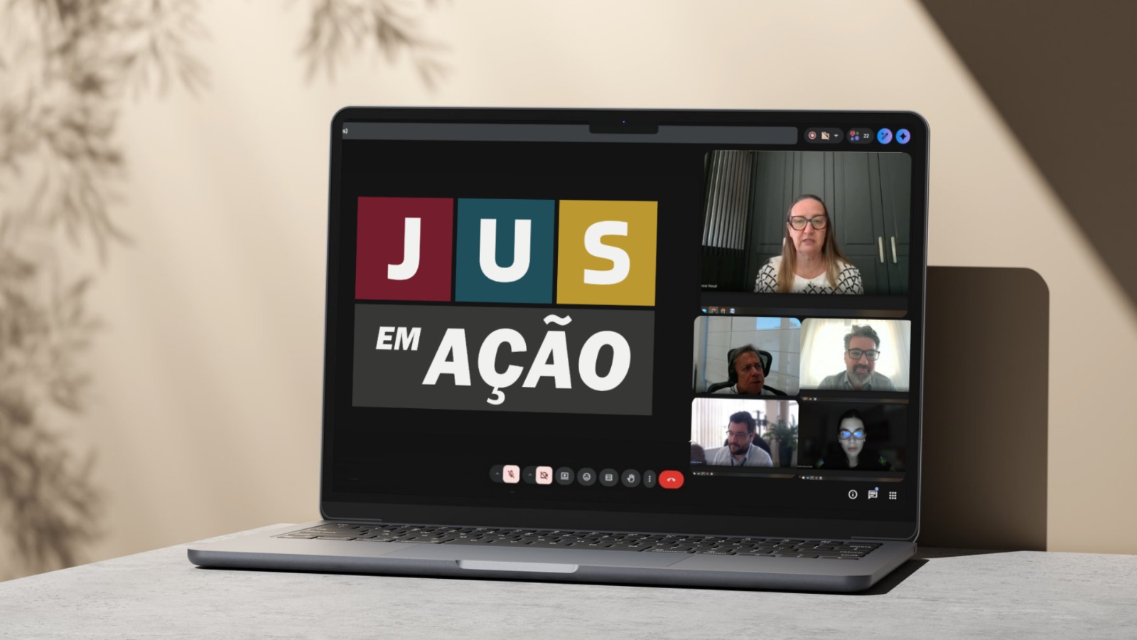 Notebook aberto sobre uma mesa exibe uma reunião virtual. Na tela, aparece o logotipo “JUS em Ação” em destaque, ao lado de janelas com participantes em videoconferência. Há pelo menos cinco pessoas conectadas, com câmeras abertas, em diferentes ambientes.