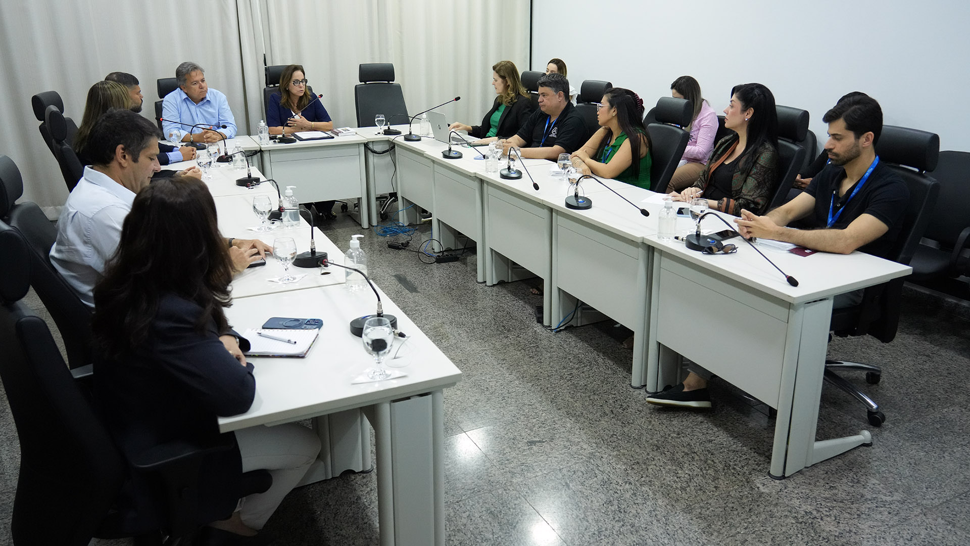 A imagem mostra uma reunião institucional na sala do Cinugep, com participantes sentados ao redor de uma mesa em formato de “U”, composta por módulos brancos e cadeiras executivas pretas. Ao todo, há cerca de dez pessoas, entre homens e mulheres, vestindo trajes formais ou sociais.  Cada lugar conta com microfone individual, além de copos com água, garrafas e materiais de apoio, como cadernos, celulares e um notebook aberto sobre a mesa. No centro da cena, uma mulher conduz a fala, enquanto os demais acompanham atentamente, alguns fazendo anotações.