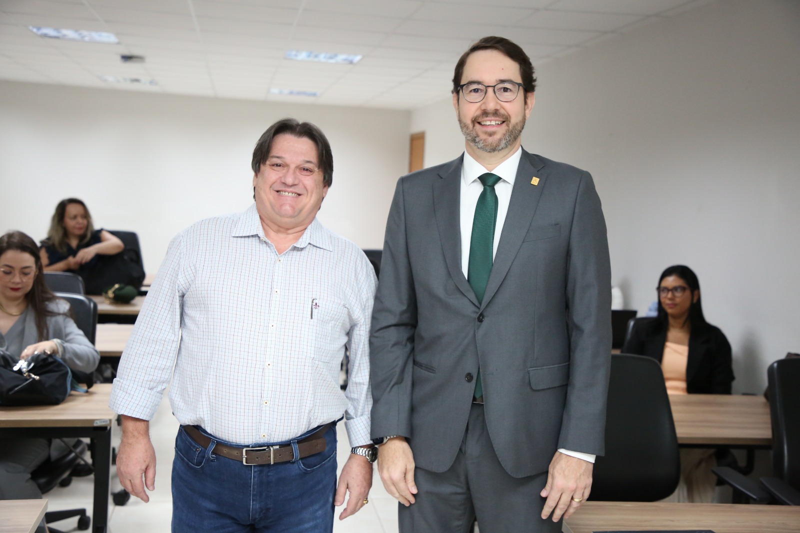 A imagem mostra dois participantes posando lado a lado durante capacitação sobre decisões do Sistema Interamericano, em ambiente institucional, sorrindo para a câmera. Ao fundo, outras pessoas aparecem sentadas em mesas, acompanhando as atividades de formação e aprendizado.