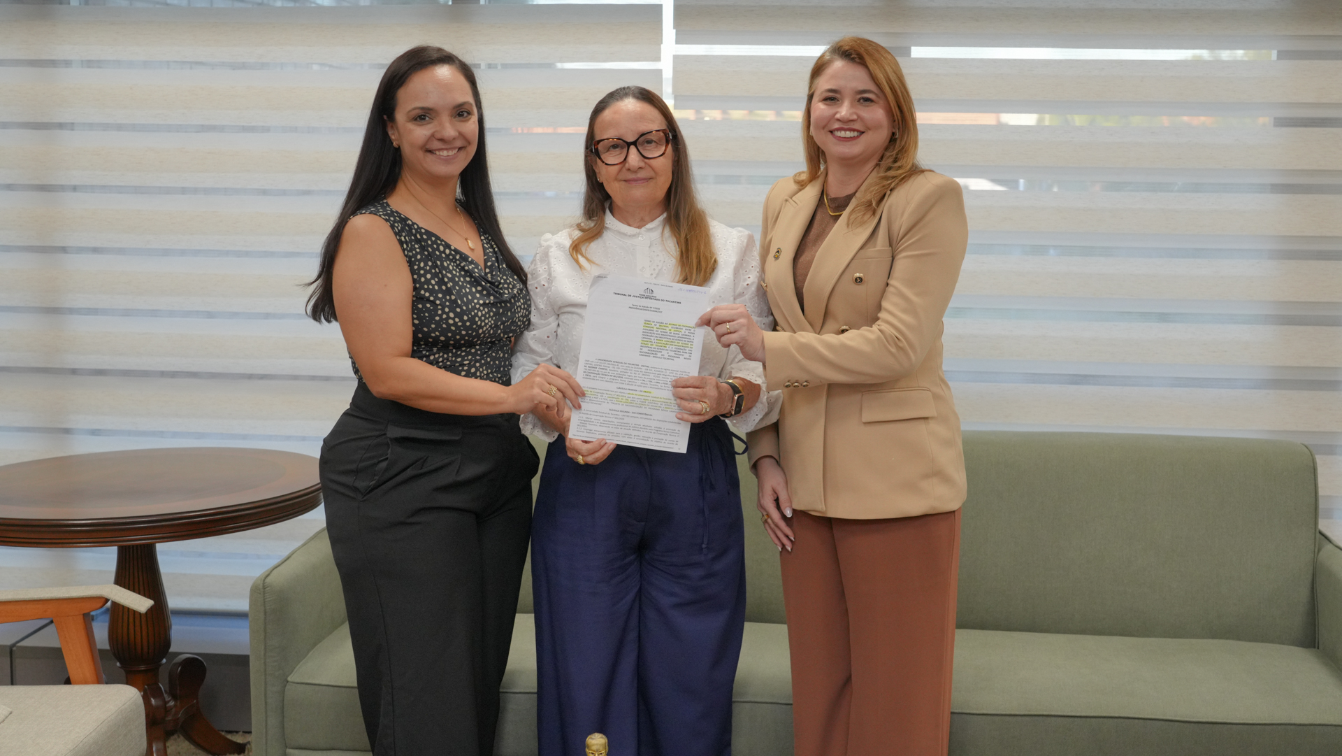 A presidente do Tribunal de Justiça do Tocantins (TJTO), desembargadora Maysa Vendramini Rosal, aparece ao centro da imagem ao lado da diretora de Comunicação do TJTO, Paula Bittencourt, à esquerda, e da vice-reitora da Unitins, à direita. As três posam sorrindo em ambiente institucional, enquanto seguram o documento que formaliza a parceria entre as instituições.