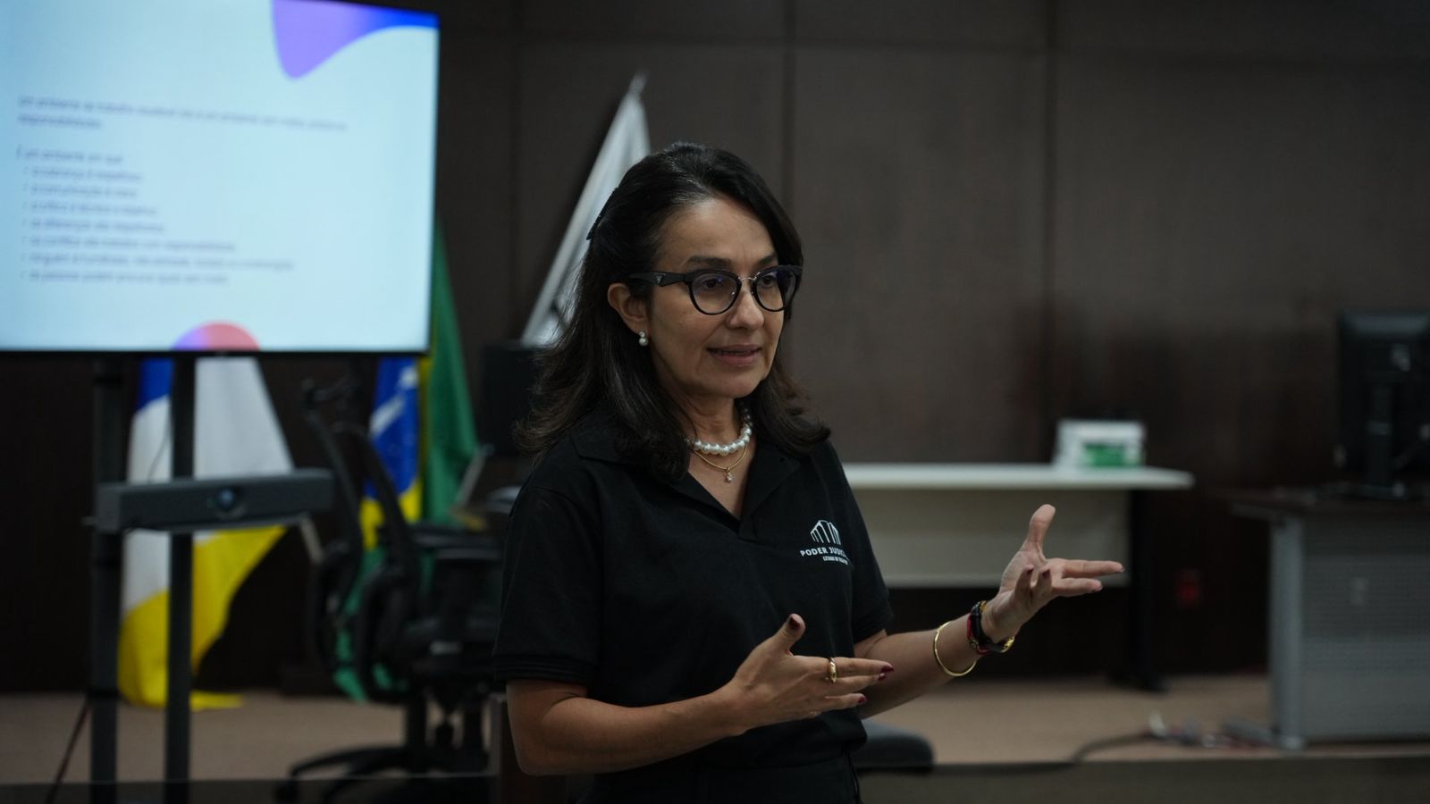 A desembargadora Hélvia Túlia Sandes Pedreira aparece em pé, durante roda de conversa promovida pelo TJTO sobre prevenção ao assédio e à discriminação no ambiente de trabalho. Vestida com camisa preta institucional do Poder Judiciário, ela gesticula enquanto conduz a apresentação. Ao fundo, um telão exibe conteúdo da palestra e bandeiras oficiais compõem o cenário do auditório.