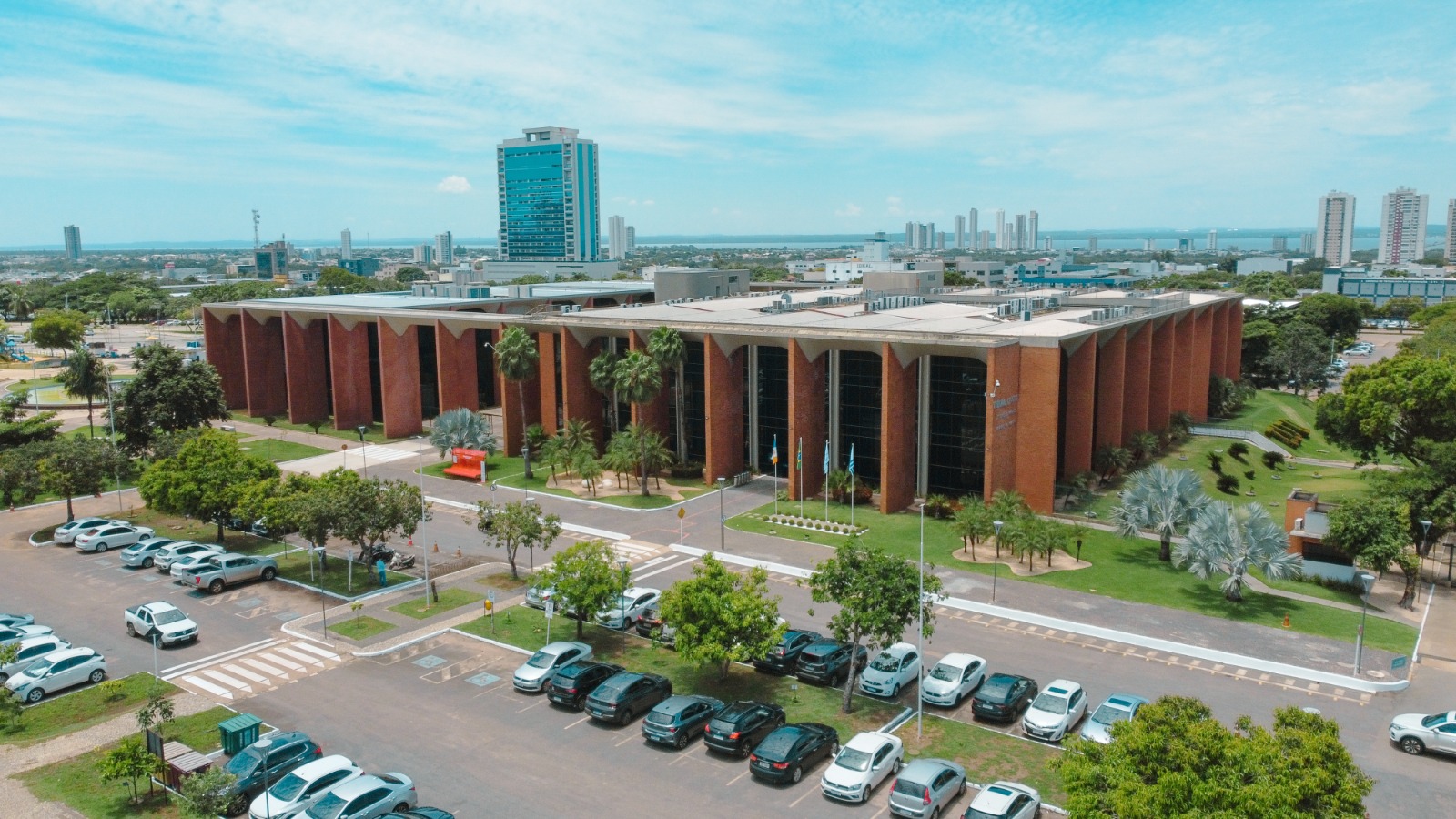 A imagem mostra vista aérea do prédio do Tribunal de Justiça do Tocantins, com arquitetura imponente, fachada em tons de vermelho com tijolinhos à vista e vidros espelhados, cercado por jardins e estacionamento. Ao fundo, aparecem edifícios da área urbana e a paisagem da cidade sob céu azul com poucas nuvens.