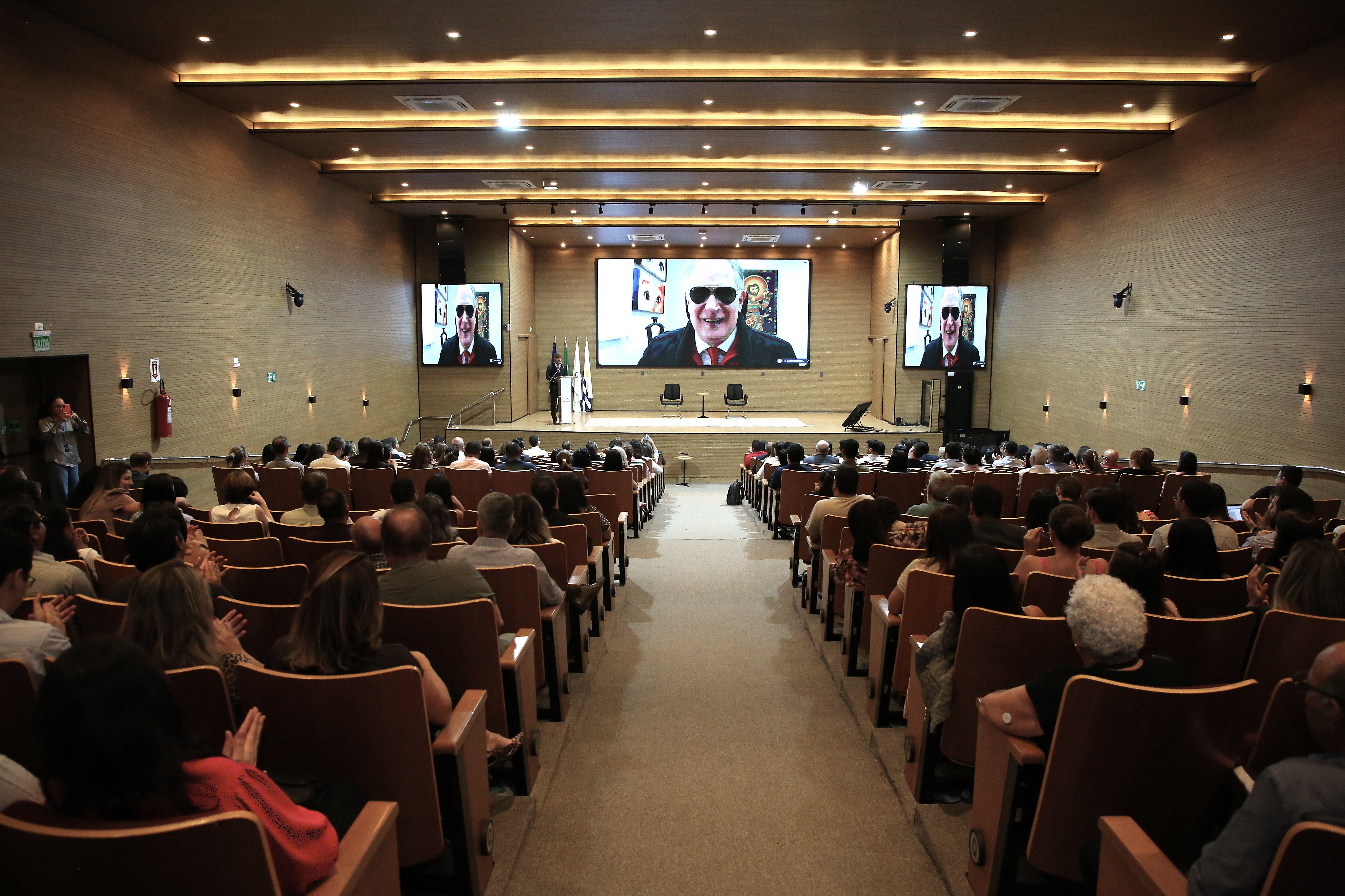 A imagem mostra auditório do TJTO com magistrados(as), servidores(as) e convidados durante a abertura do curso Boas práticas de acessibilidade no Poder Judiciário, realizado pelo TJTO. No telão, o desembargador Ricardo Tadeu Marques da Fonseca participa de forma online como palestrante da abertura, compartilhando reflexões sobre acolhimento, direitos e inclusão de pessoas com deficiência.