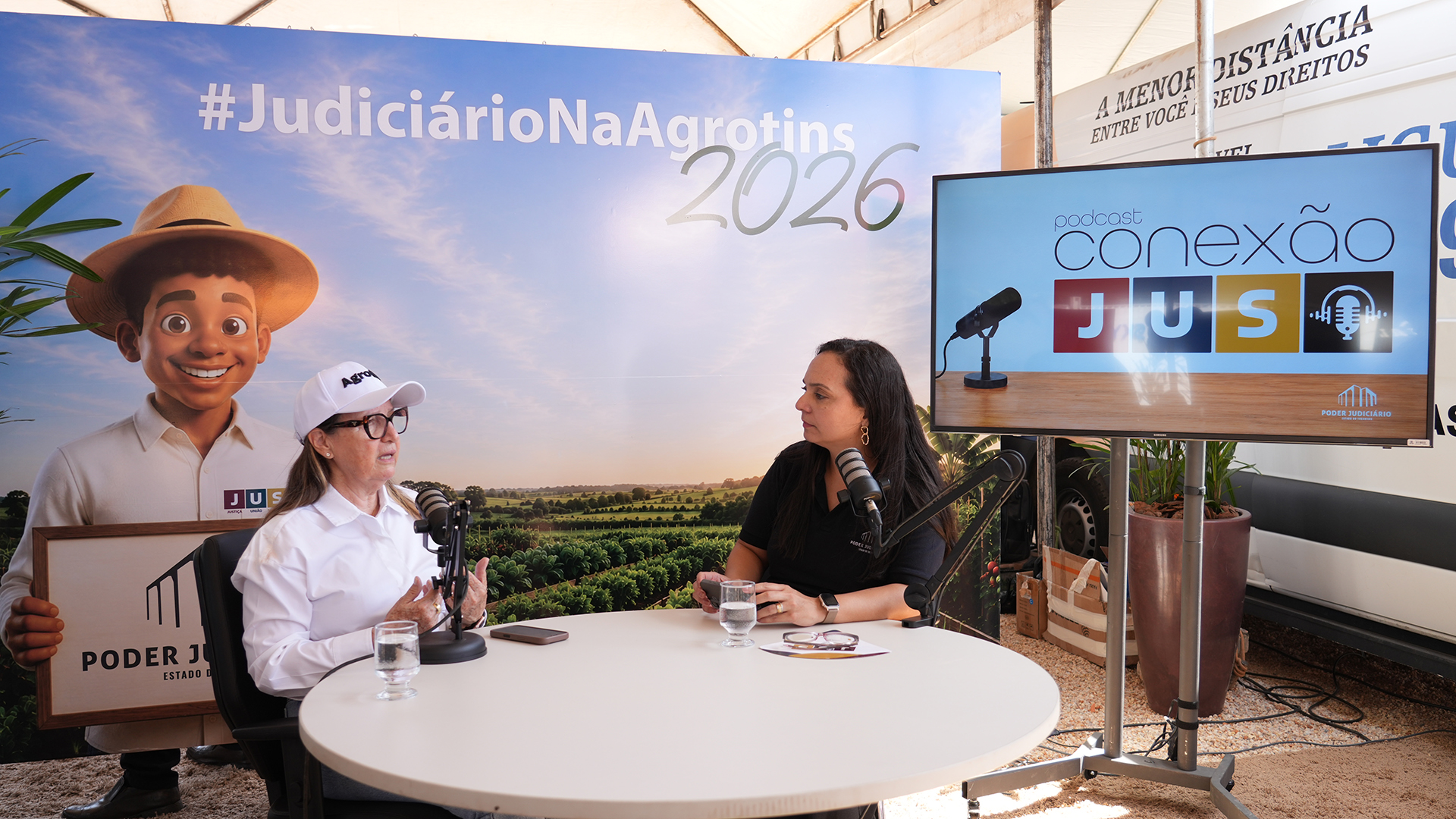 A presidente do Tribunal de Justiça do Tocantins, desembargadora Maysa Vendramini Rosal, participa da gravação do podcast “Conexão JuS” ao lado da jornalista Paula Bittencourt, diretora do Centro de Comunicação Social do TJTO, no estande do Poder Judiciário durante a Agrotins 2026. O ambiente conta com estrutura de gravação profissional, painel temático da feira e monitor com a identidade visual do podcast.