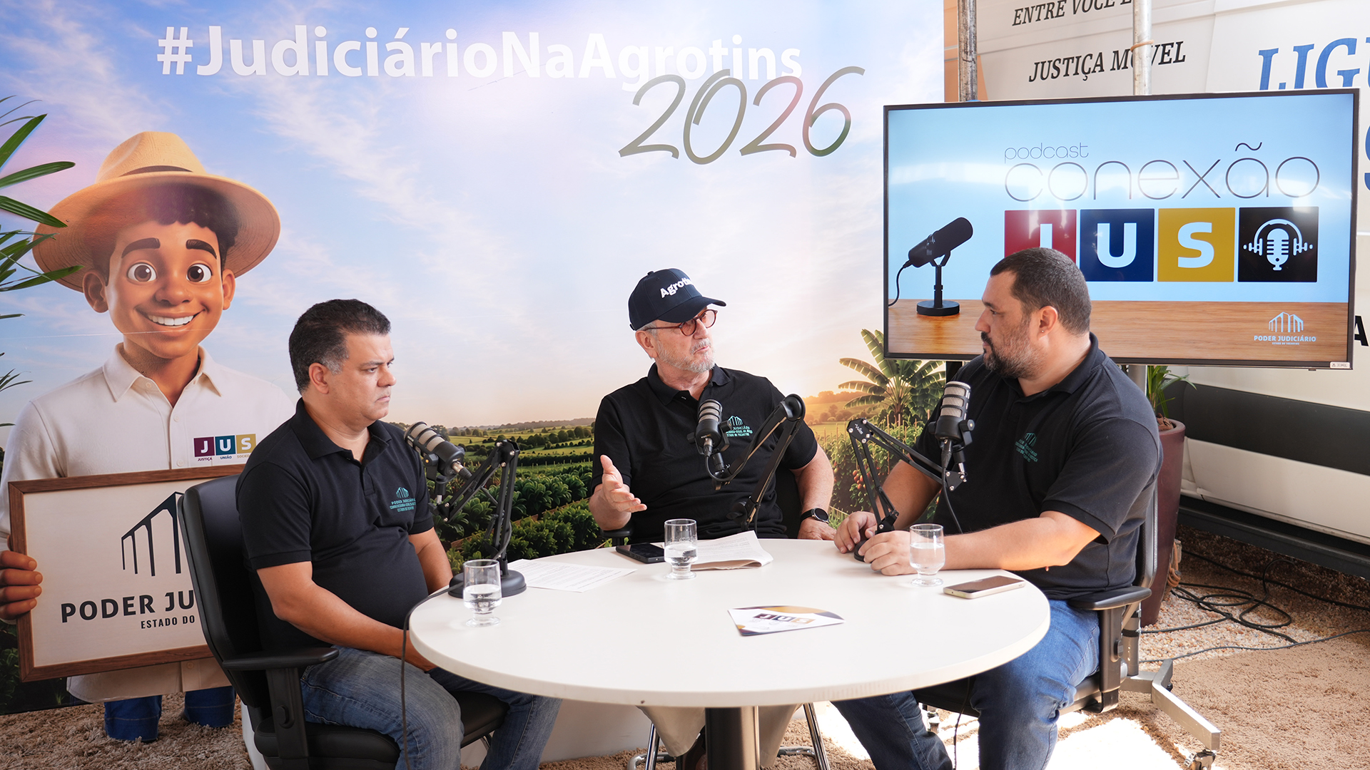 O desembargador Pedro Nelson de Miranda Coutinho, ao centro, e o juiz Jordan Jardim, à esquerda, participam de gravação do podcast “Conexão JuS” no estande do Poder Judiciário do Tocantins durante a Agrotins 2026. O episódio abordou regularização fundiária e segurança jurídica no campo, com mediação do jornalista Philipe Bastos.