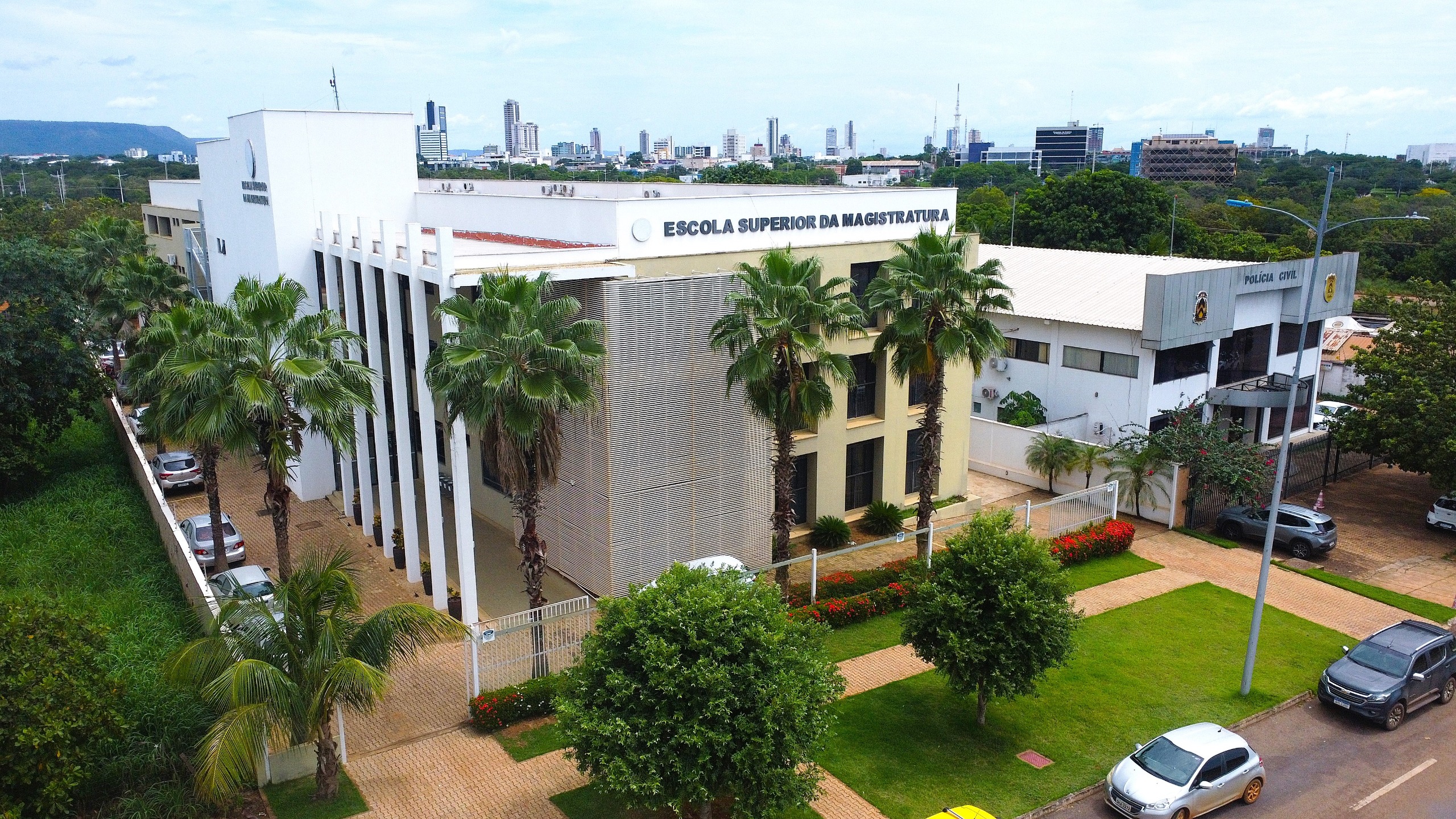 Imagem aérea da fachada da Escola Superior da Magistratura Tocantinense (Esmat), em Palmas. O prédio de arquitetura moderna aparece cercado por palmeiras, jardins e área gramada. Ao lado da unidade, também é possível ver a sede da Polícia Civil. Ao fundo, aparece parte da paisagem urbana da capital tocantinense.